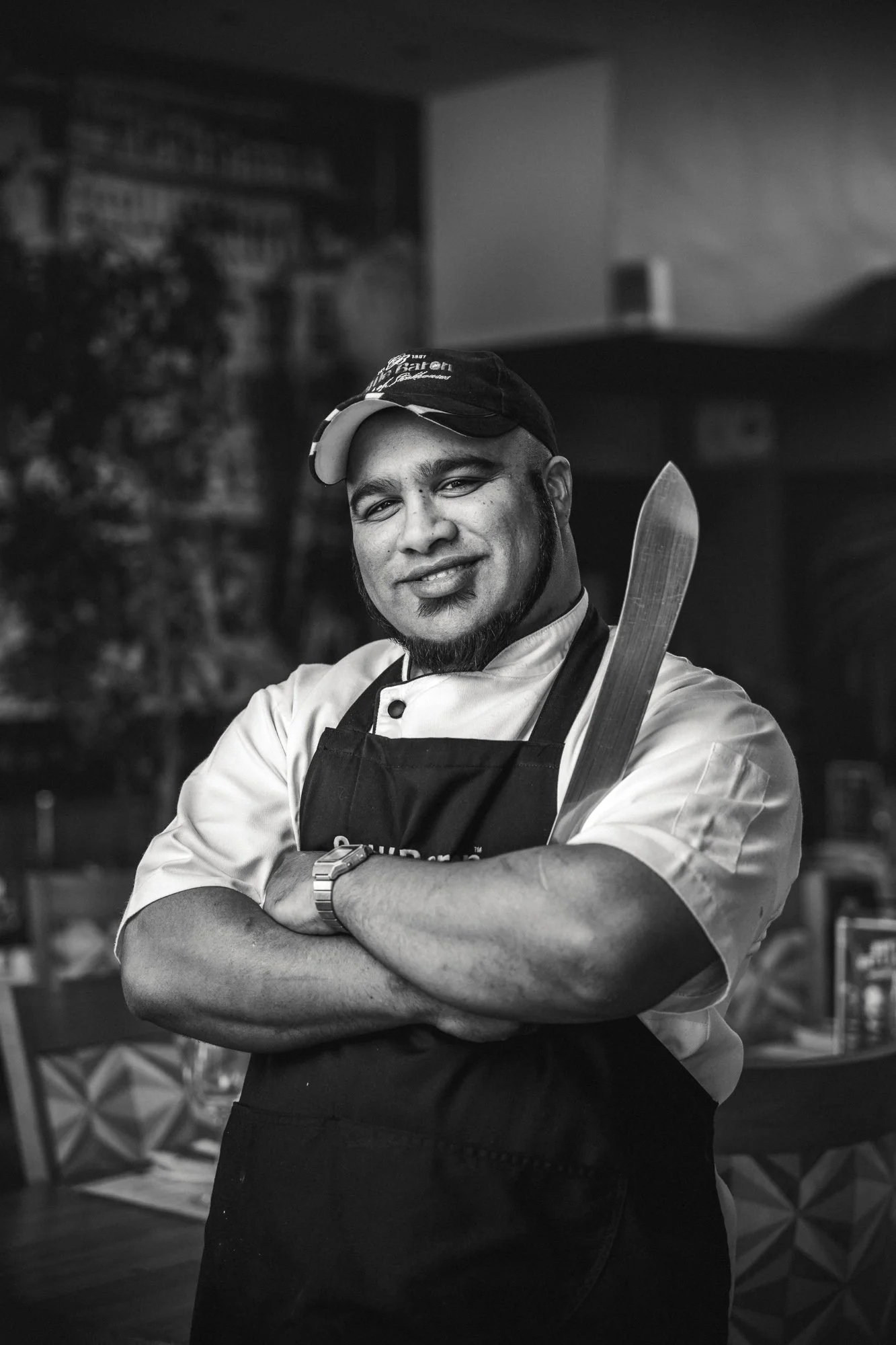 A man wearing a baseball cap, chef's coat, and apron smiling with crossed arms, holding a knife, in a kitchen setting.