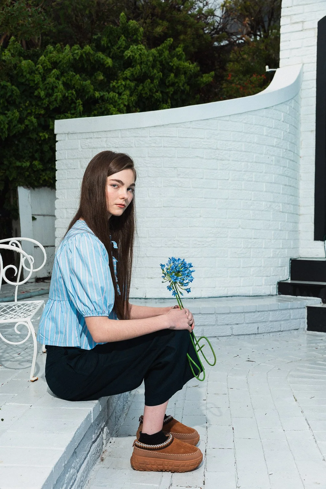 A young woman with long brown hair, wearing a blue and white striped top, black pants, and brown shoes, seated outdoors on a white brick ledge, holding a blue flower, with a white brick wall and green bushes in the background.