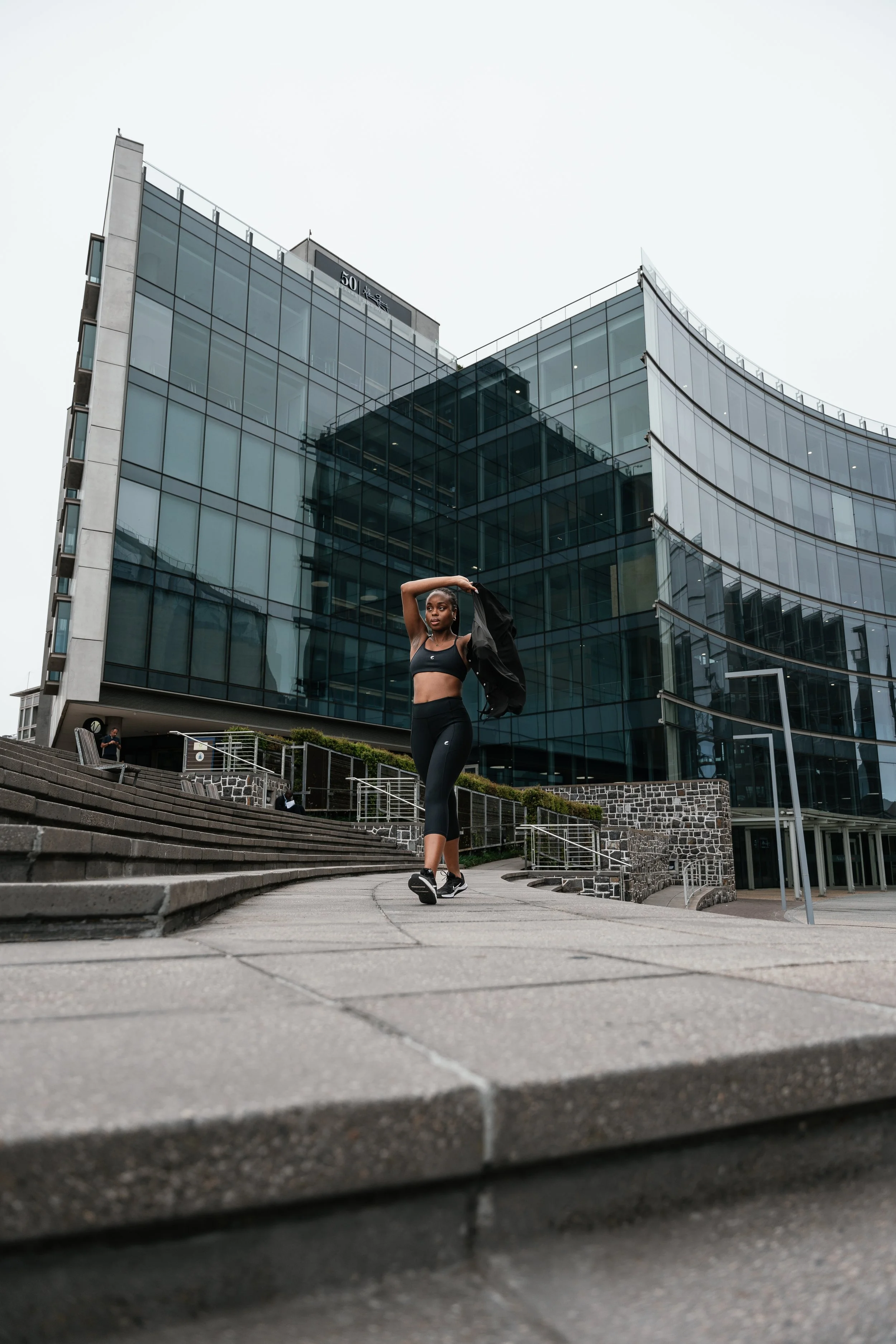 A woman in athletic wear stretching outside a modern glass office building with a curved facade, on outdoor stairs.