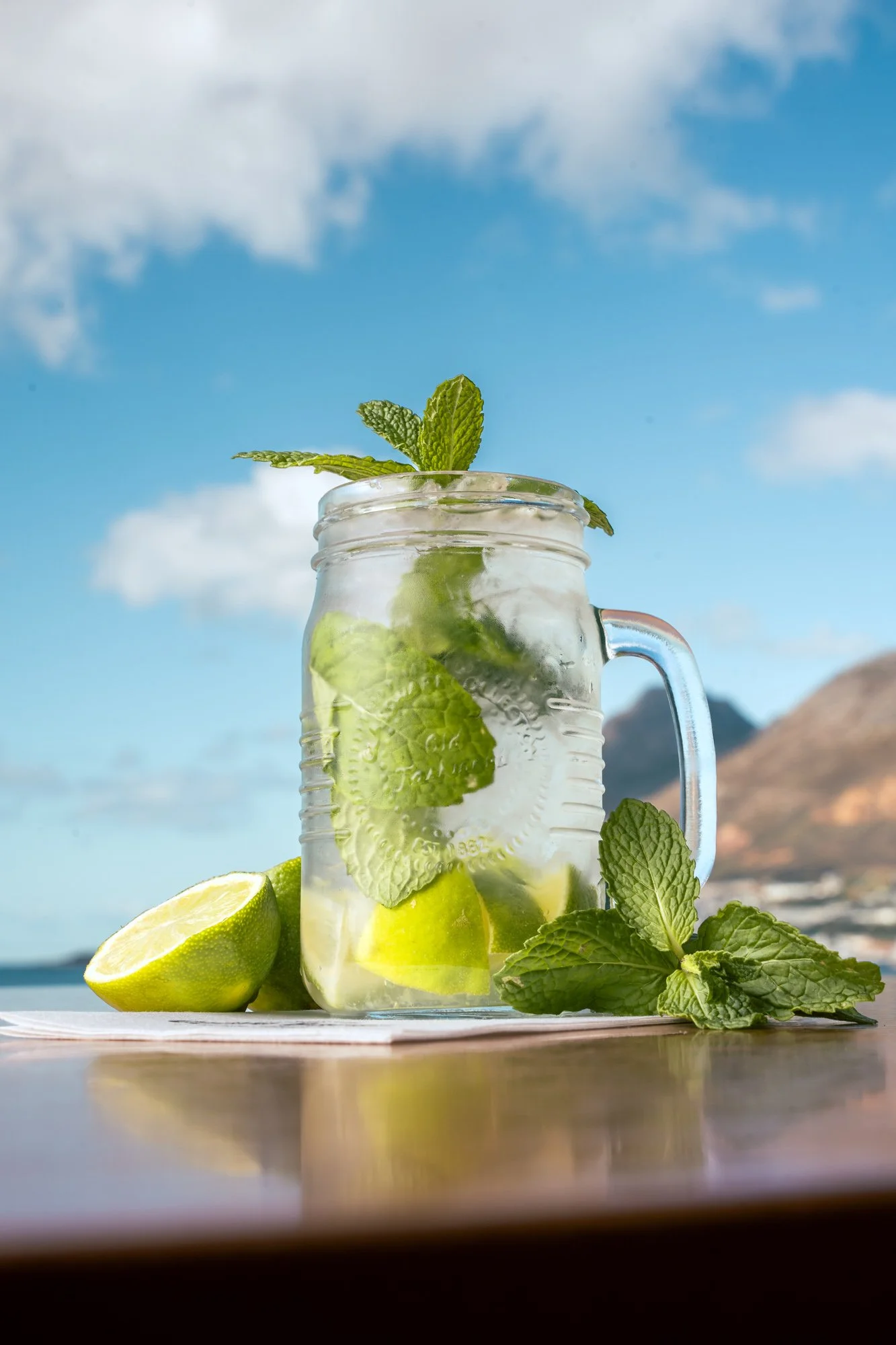 Glass jar of refreshing lemon mint infused water with lime wedges and mint leaves, outdoors under a blue sky with clouds.