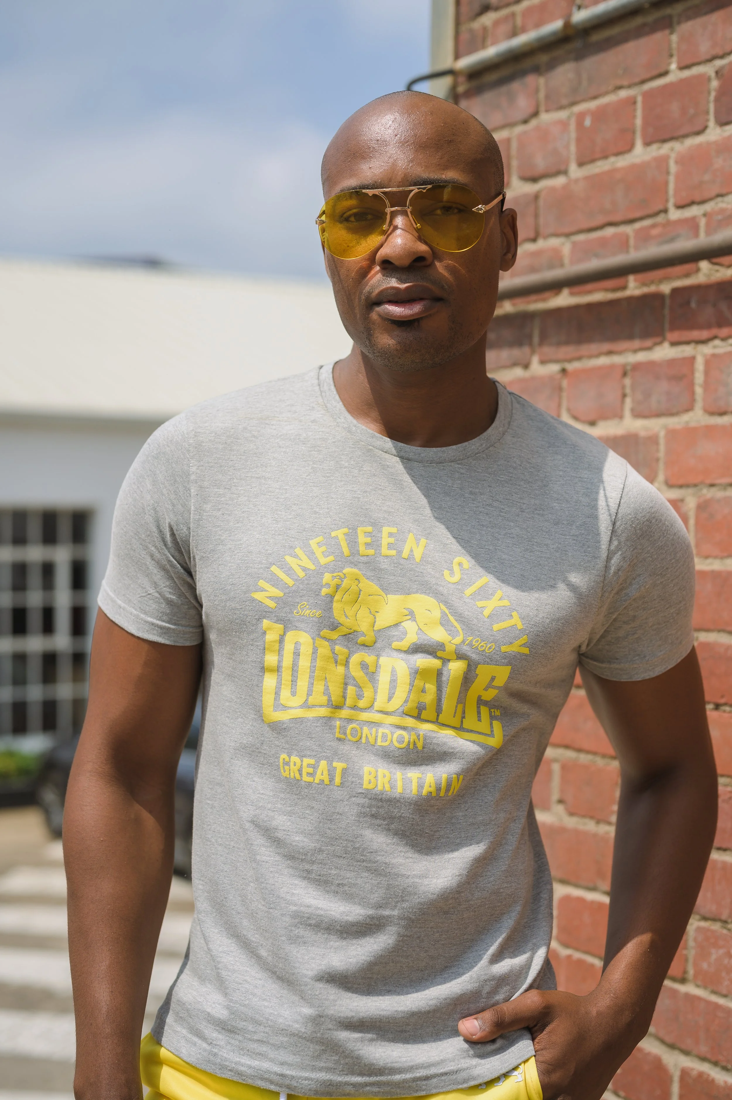 A man with a shaved head wearing yellow-tinted sunglasses and a gray T-shirt with yellow text, standing outdoors against a brick wall with a clear sky and residential buildings in the background.