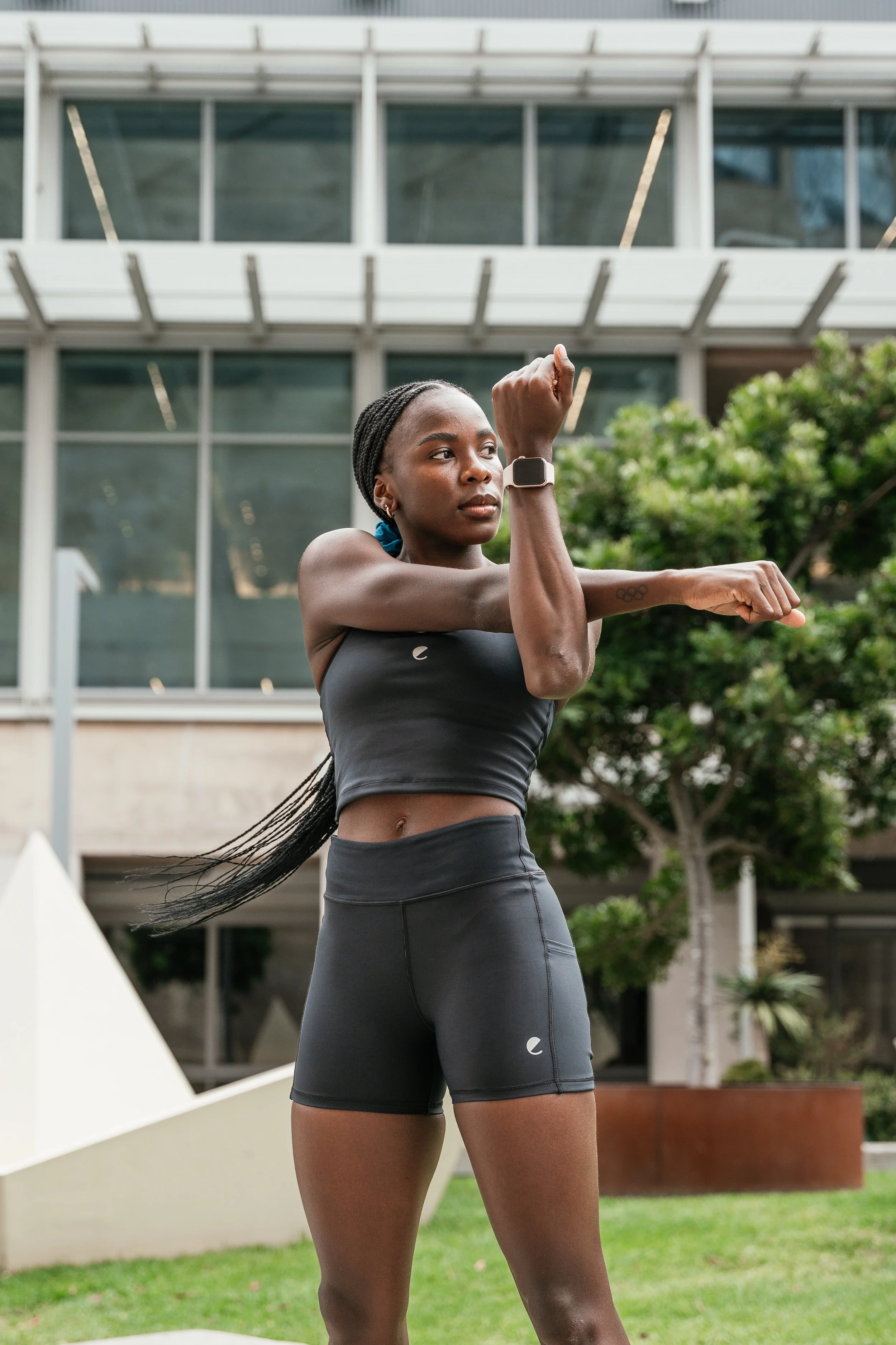 A woman doing a stretching exercise outdoors with modern building and greenery in the background.