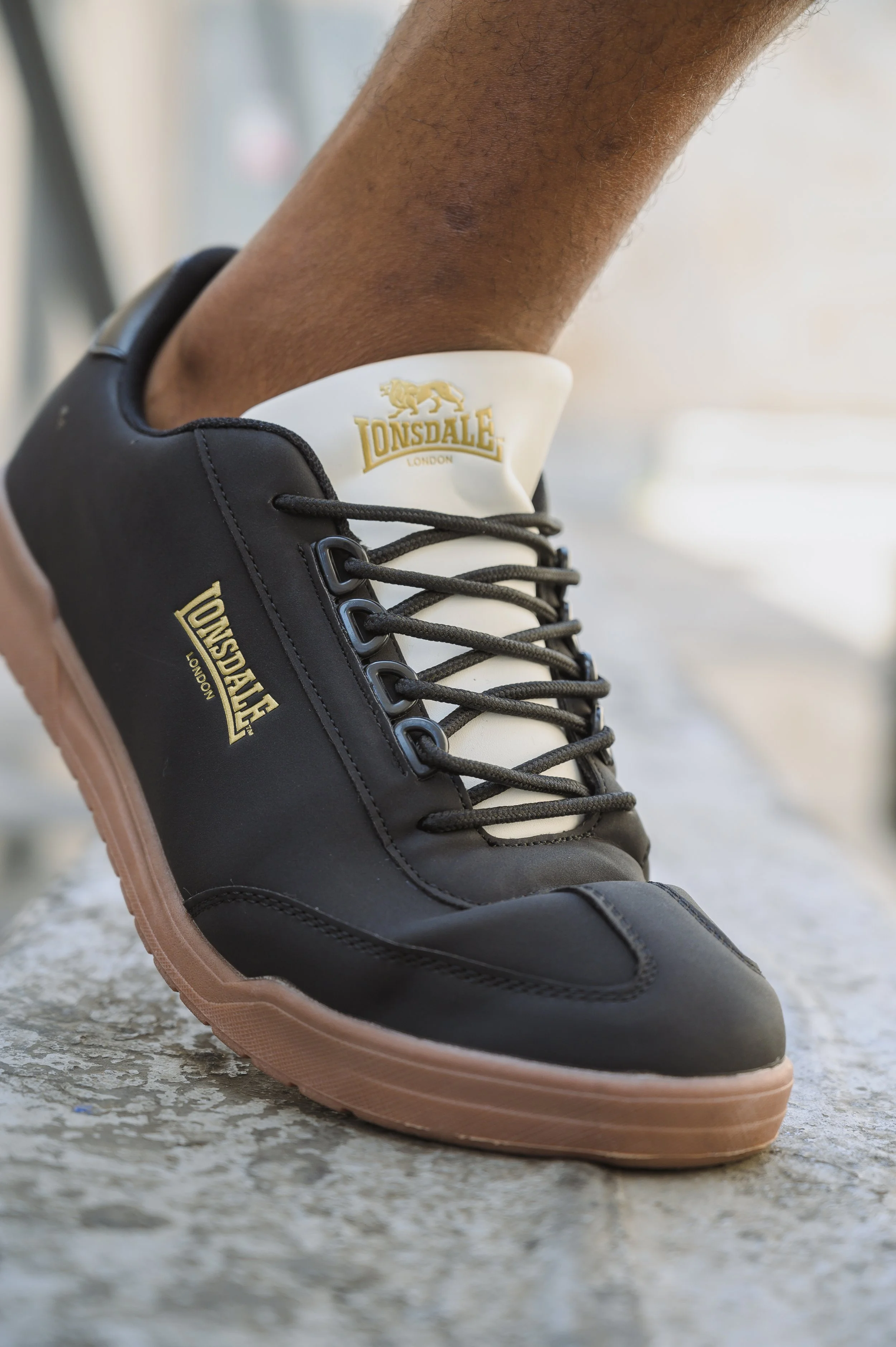 Close-up of a person's foot wearing a black and white Lonsdale London sneaker, standing on a concrete surface.