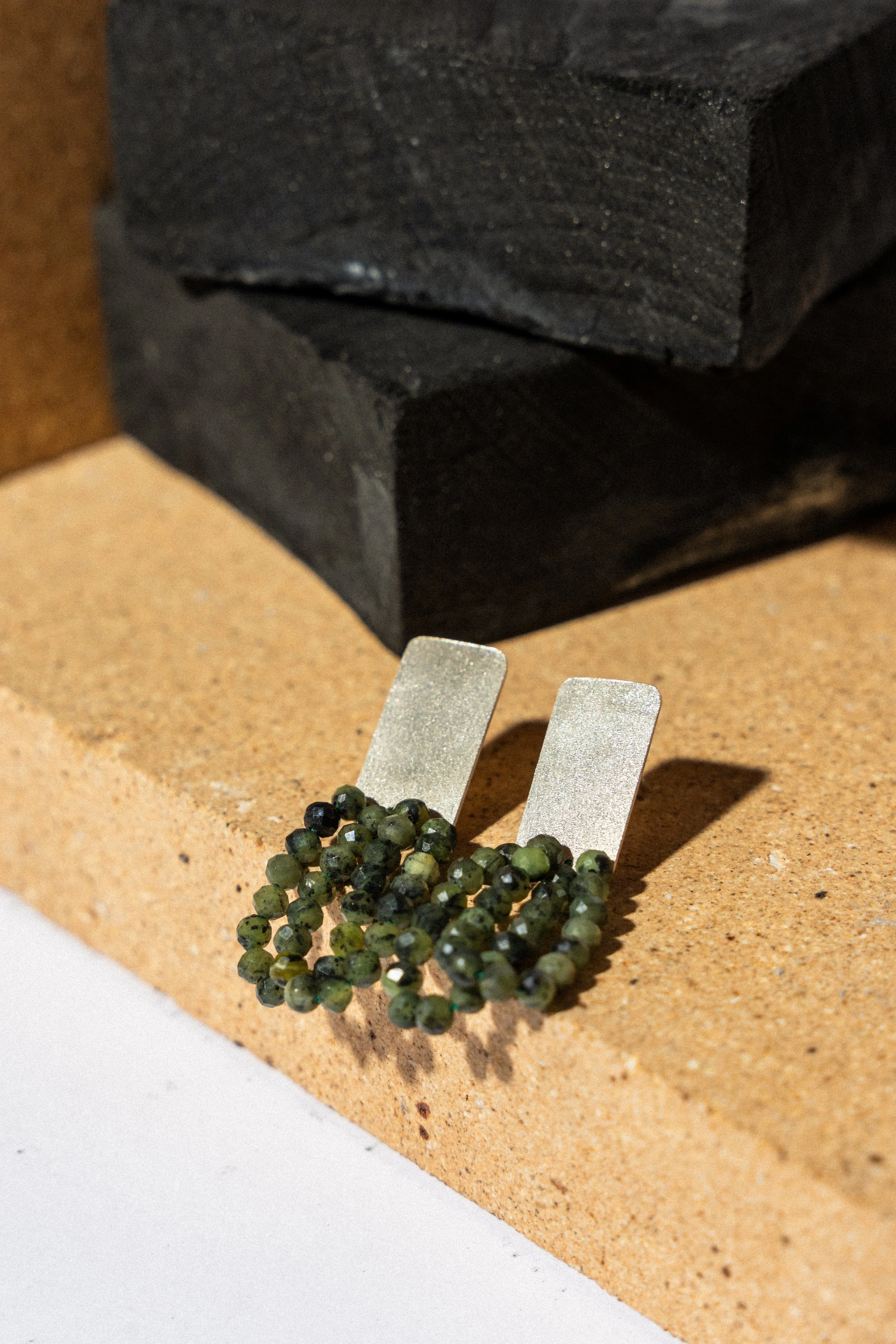 Close-up image of a small jewelry clasp attached to a string of green, black, and gray beads with metallic accents, resting on a cardboard surface.