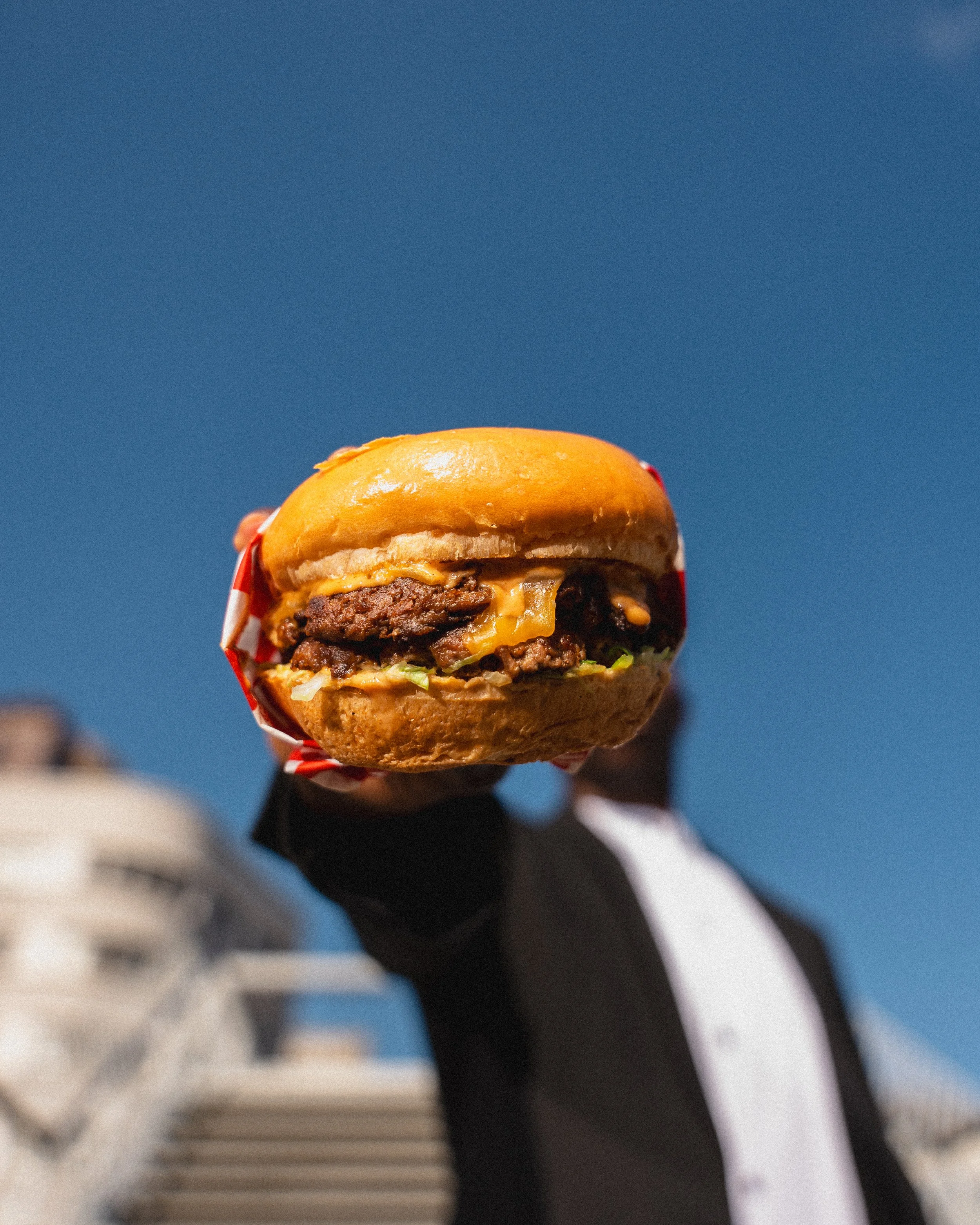 Close-up of a cheeseburger with a sesame seed bun, melted cheese, a beef patty, lettuce, and tomato against a bright blue sky.