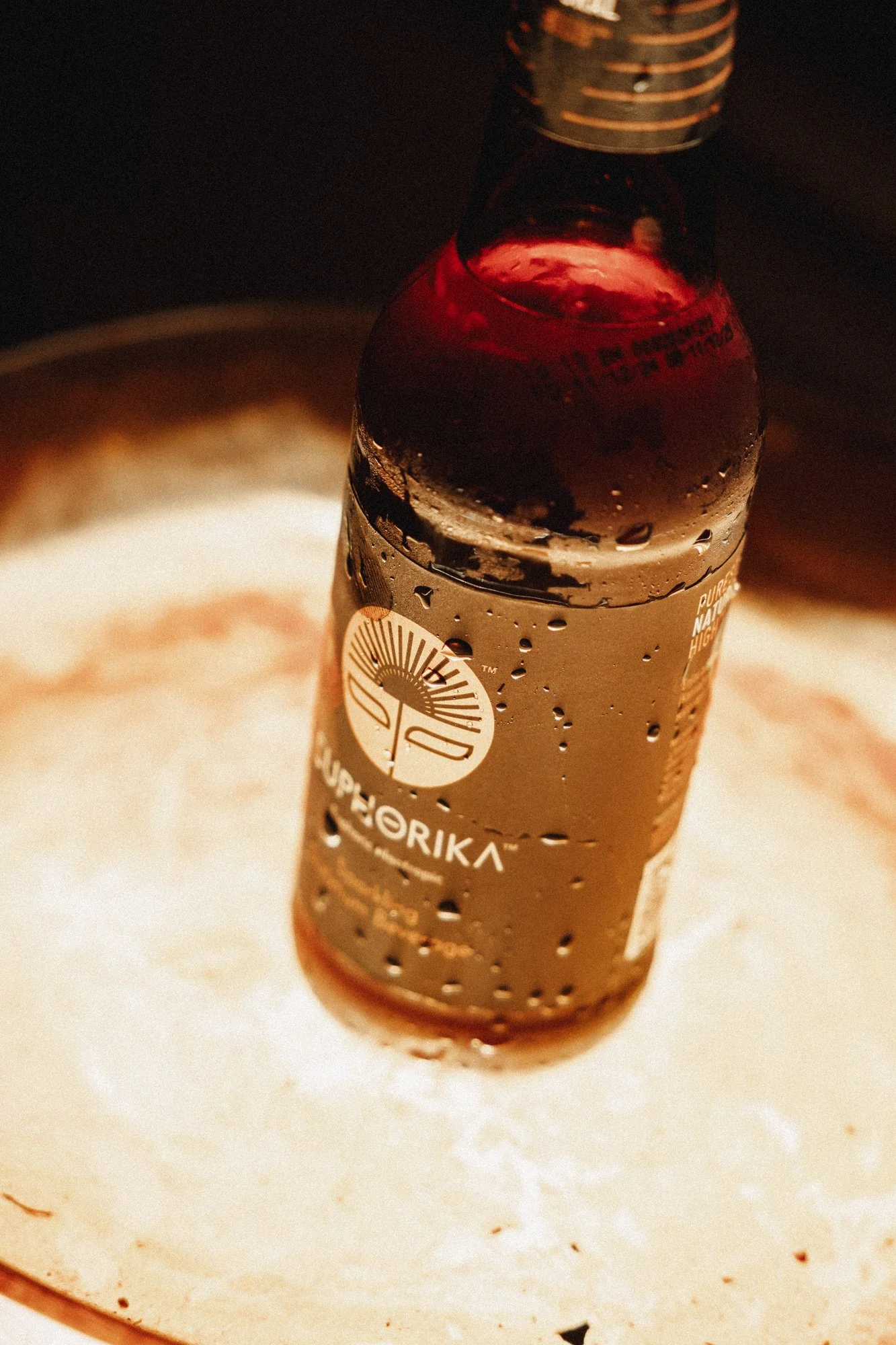 Close-up of a glass bottle with condensation, placed on a white surface. The bottle has a dark label with white and gold text and a logo. The background is dark and out of focus.