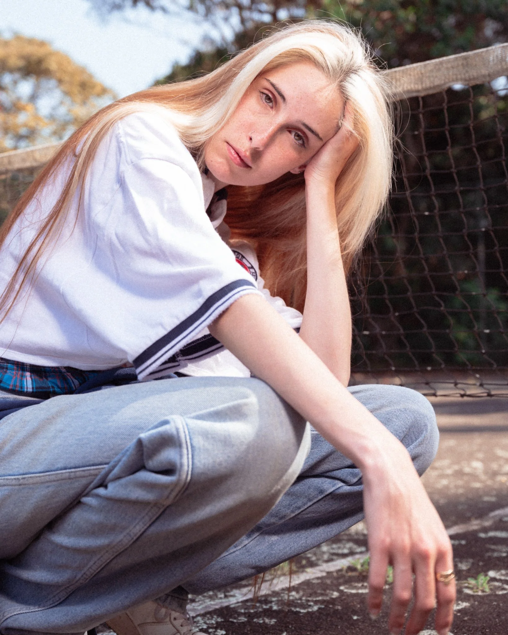 A young woman with long blond hair wearing a white shirt with a patch on the sleeve, gray jeans, and beige shoes, sitting on the ground outdoors near a tennis net, with trees and blue sky in the background.