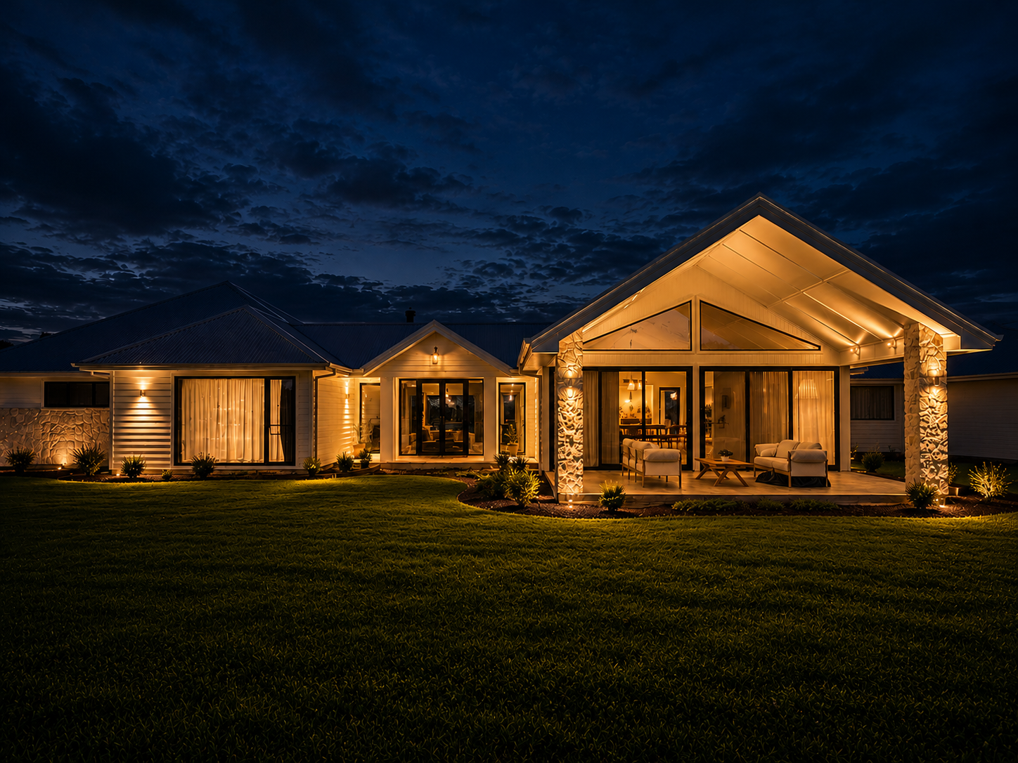 Modern residential home lit at dusk, with warm exterior facade lighting, illuminated gable ceiling, garden spotlights and glowing interior lighting throughout.