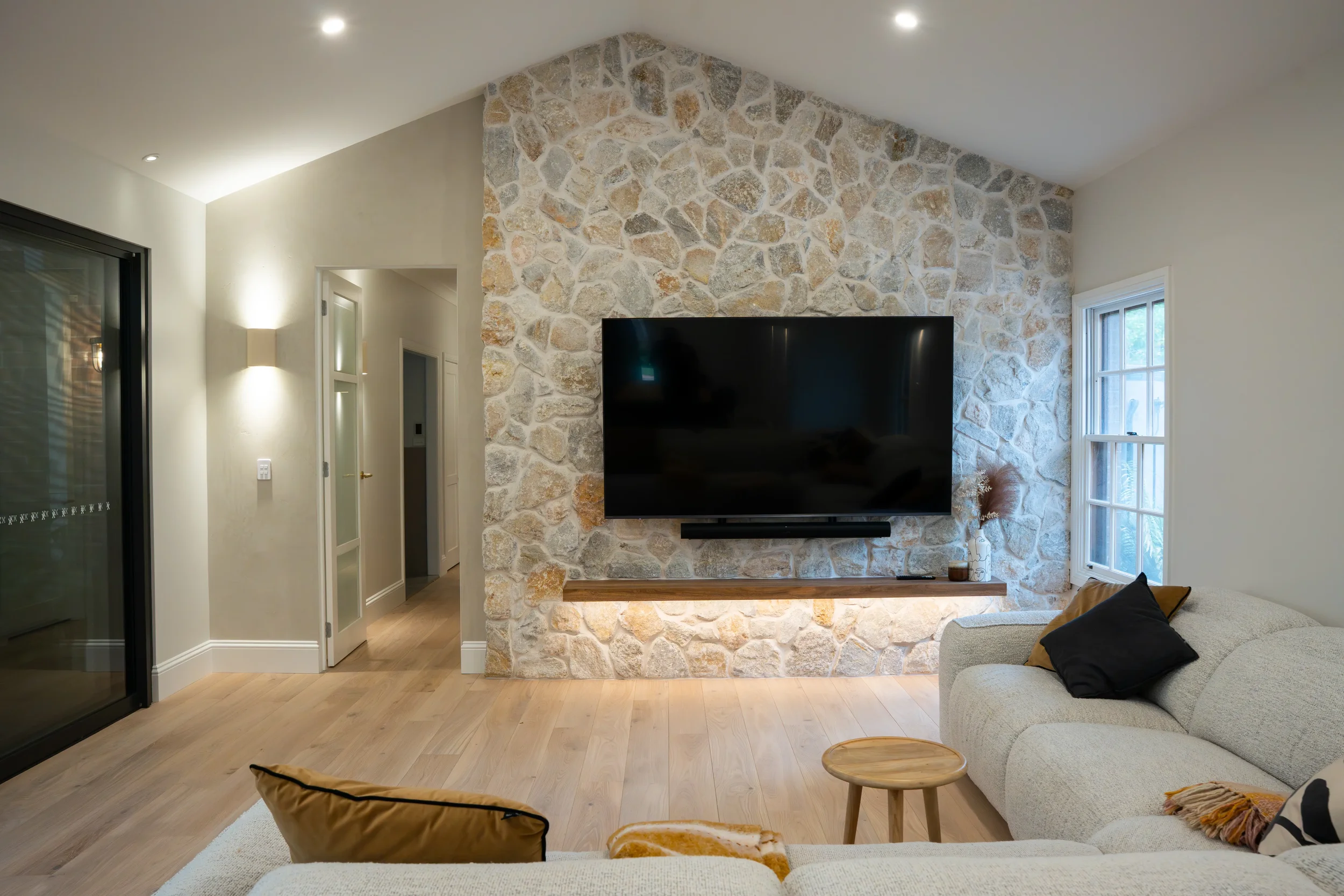 Modern living room with a wall-mounted TV and soundbar on a natural stone feature wall, concealed cabling, recessed downlights and LED strip lighting beneath a floating timber shelf.