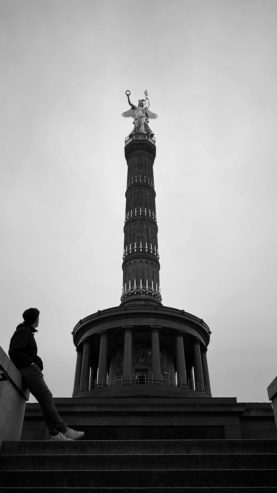Une personne assise sur une rambarde regarde une grande colonne avec une statue en haut représentant une figure féminine avec des ailes et tenant une branche dans une main et un objet dans l'autre. Le ciel est gris et nuageux.