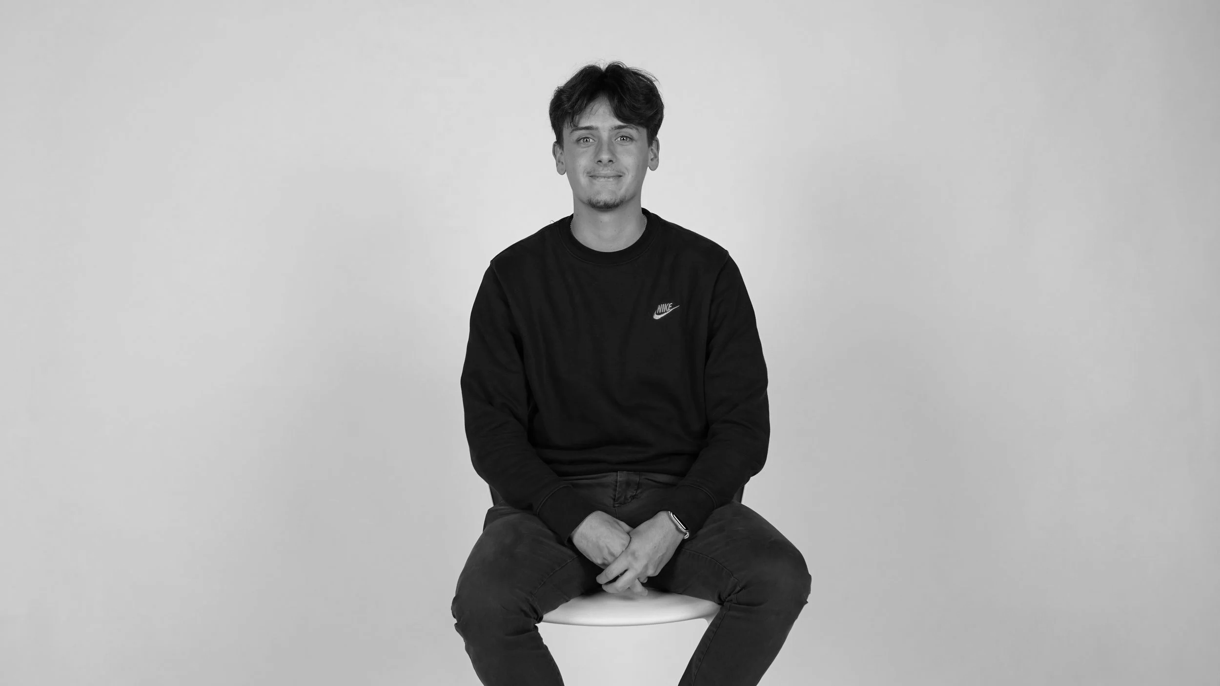 Un jeune homme assis sur un tabouret, portant un sweat à capuche noir, souriant, dans une composition en noir et blanc.