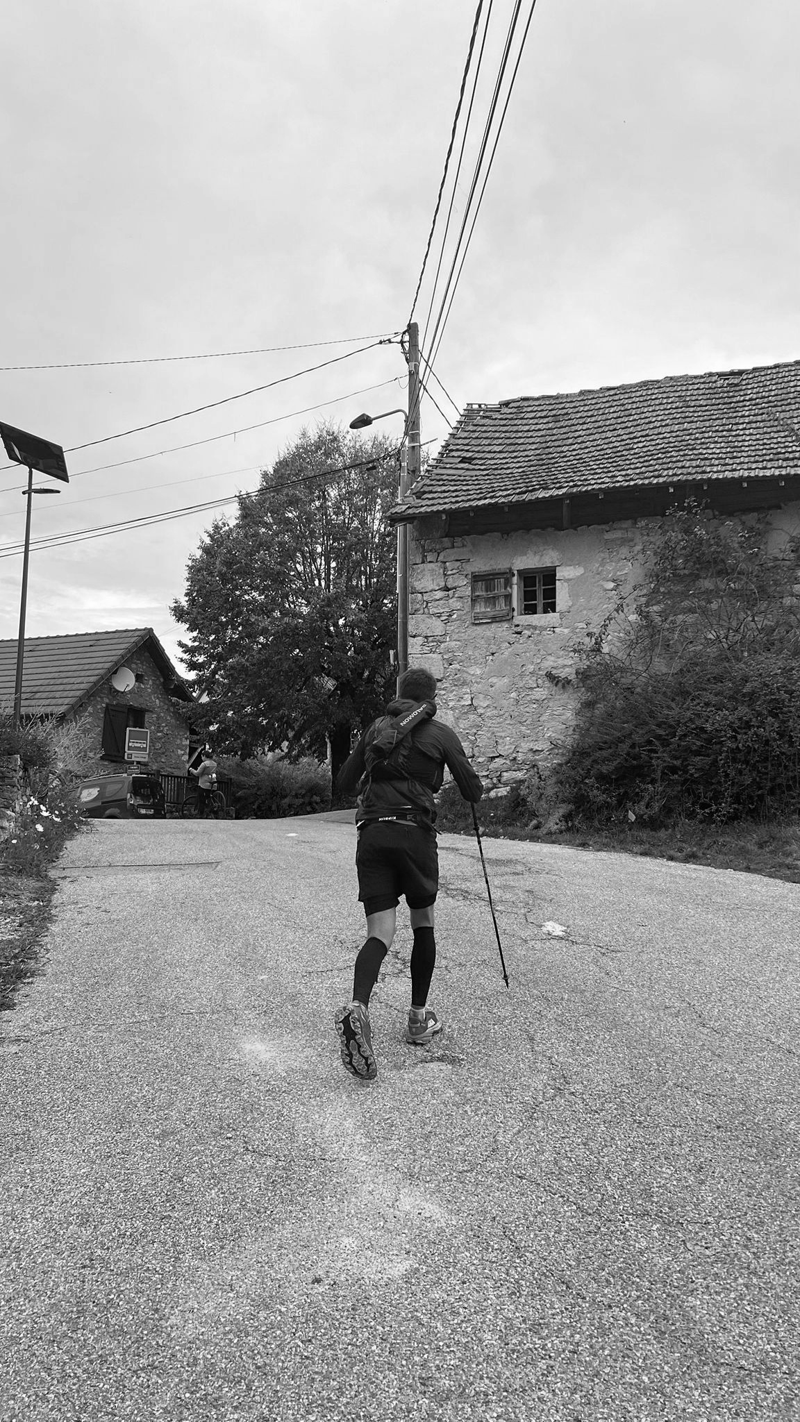 Homme en course sur une route montante dans un village avec des maisons en pierre et des arbres, ciel nuageux, en noir et blanc.