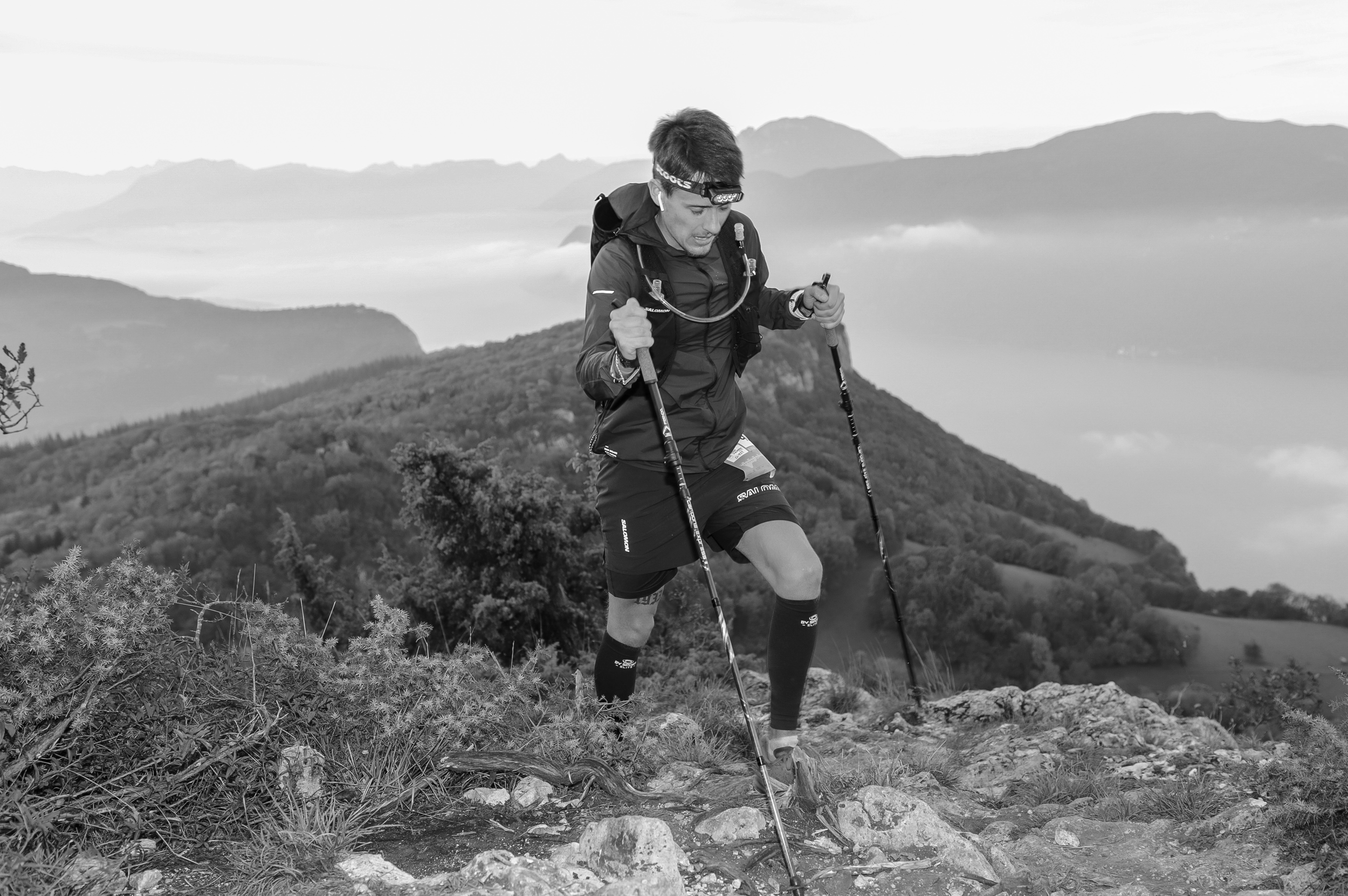 Un homme en vêtements de course avec des bâtons de marche, gravissant une montagne avec un paysage de montagnes en arrière-plan.
