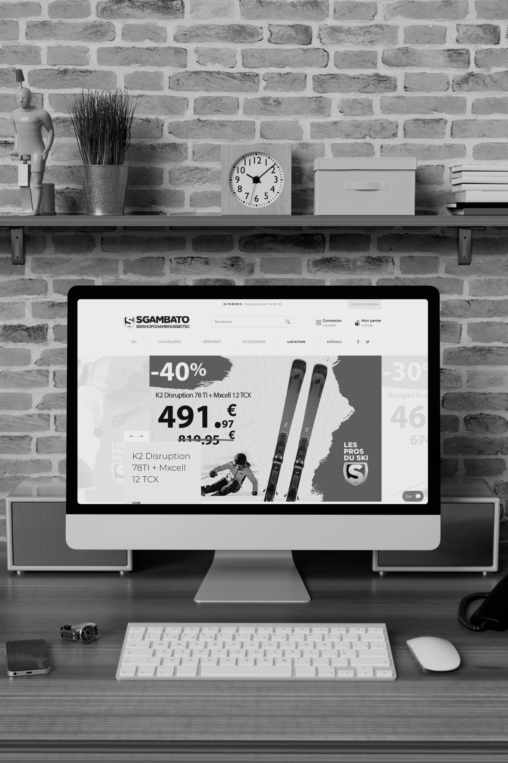 Écran d'ordinateur affichant une publicité pour des skis à prix réduit, avec un fond en bois, une souris, un clavier, deux haut-parleurs, une montre, un téléphone portable, un téléphone fixe et une étagère avec des objets décoratifs et un horloge.