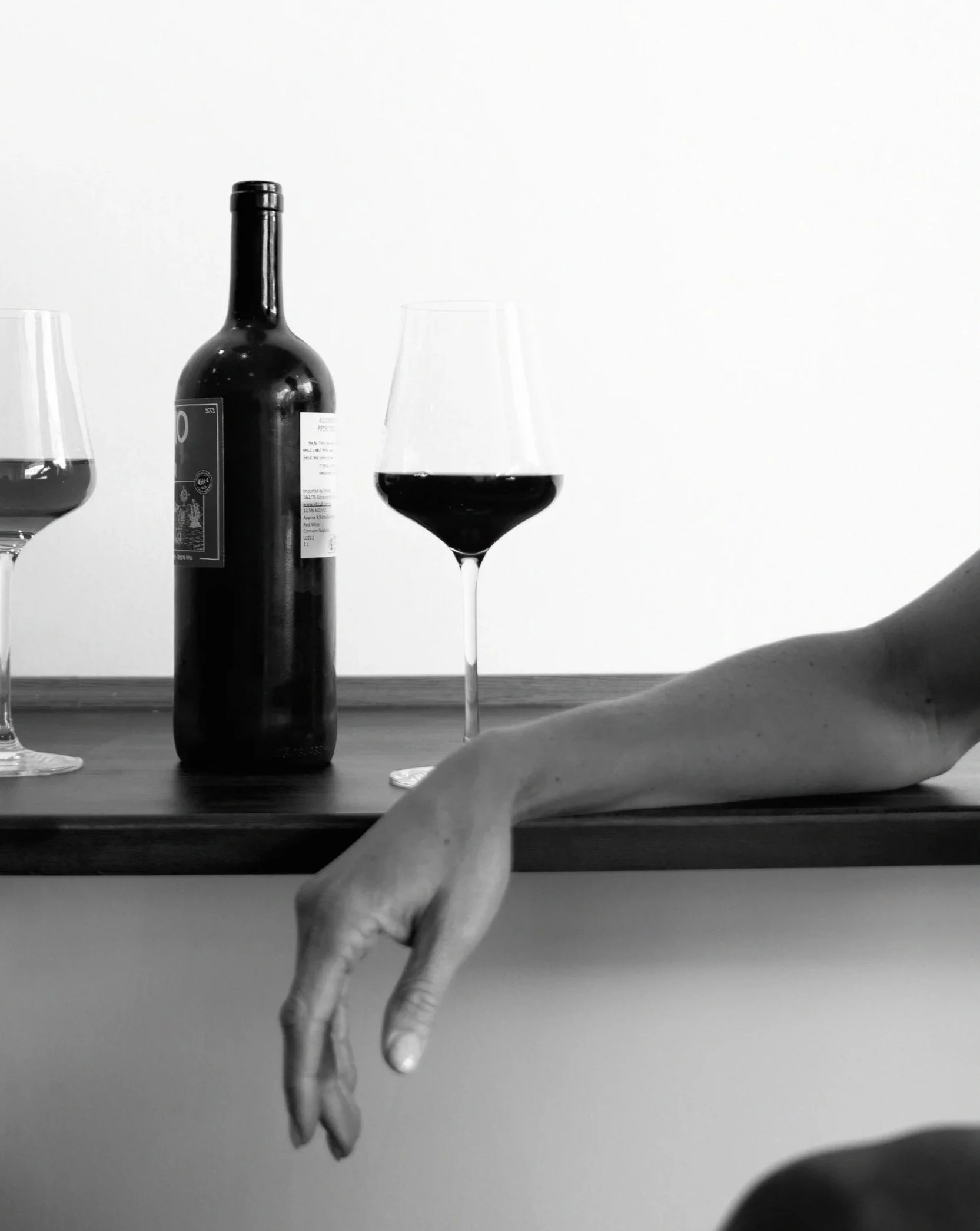 A black wine bottle and two wine glasses filled with red wine on a wooden table, with a person's hand visible in the foreground.