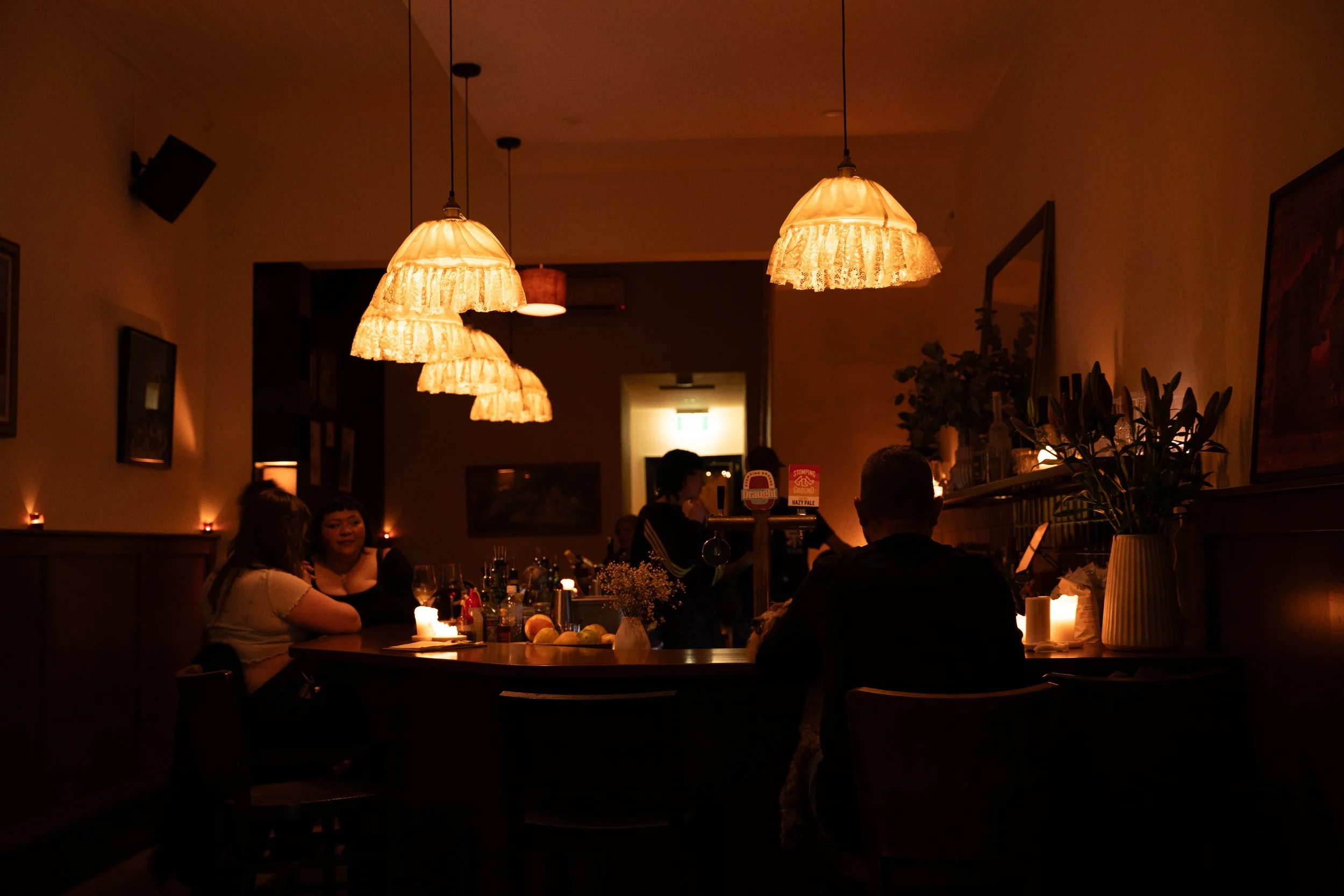 Bernie'sFlintaBar A dimly lit restaurant scene with hanging pendant lights, people sitting at a table with candles, flowers, and drinks, and wall art, creating a cozy atmosphere.