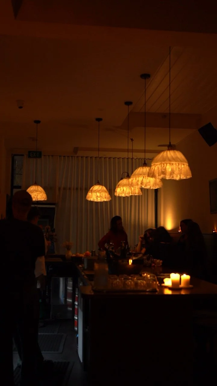 Dimly lit restaurant or bar with hanging lamps and candles on the table, with people sitting and standing around.