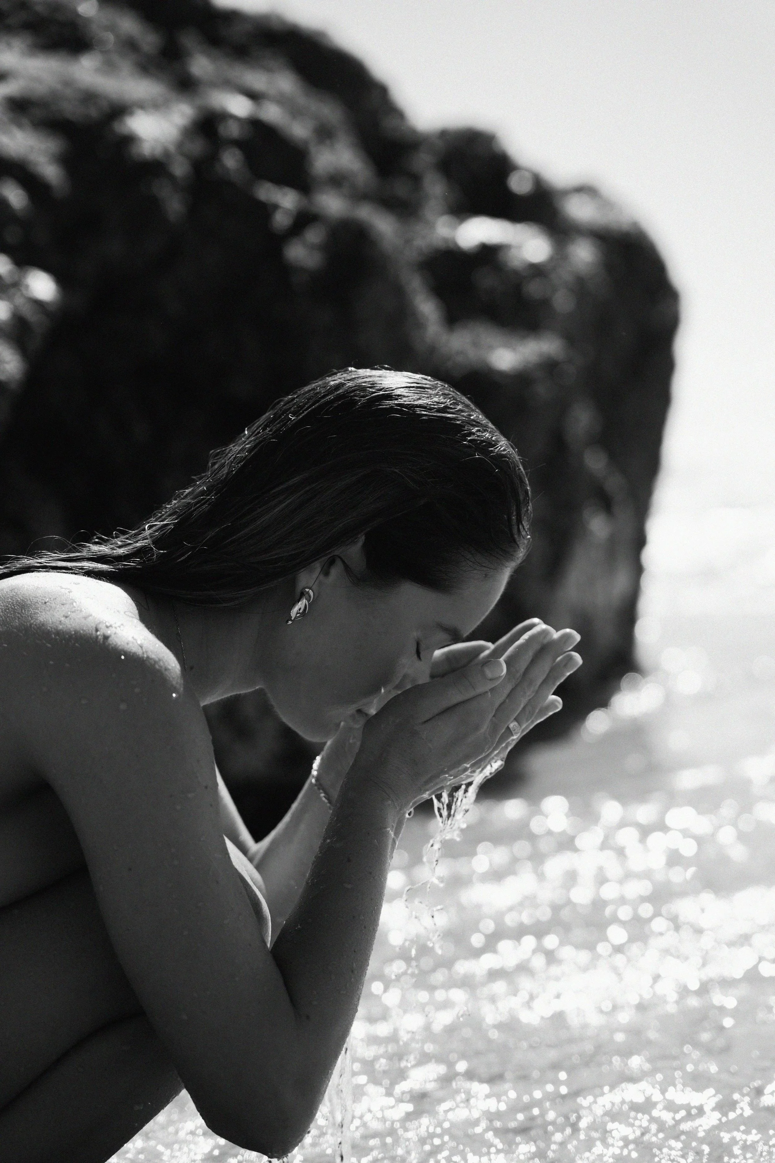 Brand campaign shoot for Melbourne small business  A woman with wet hair and a necklace, with her hands in a prayer position near her face, appears to be at the beach, with the ocean and sunlight reflected on the water in the background.