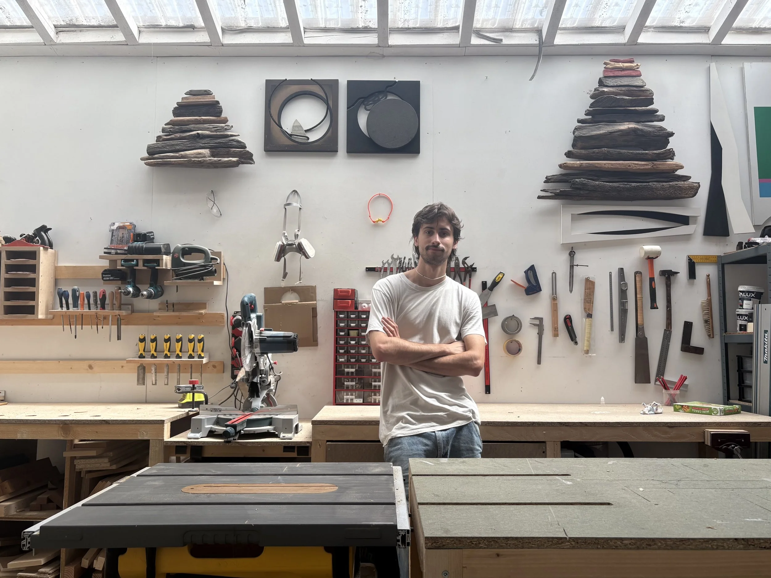 Un homme se tient dans un atelier de menuiserie, entouré d'outils suspendus au mur et posés sur un établi, sous un toit en verre, avec deux sculptures en bois en forme de pyramide dans le fond.