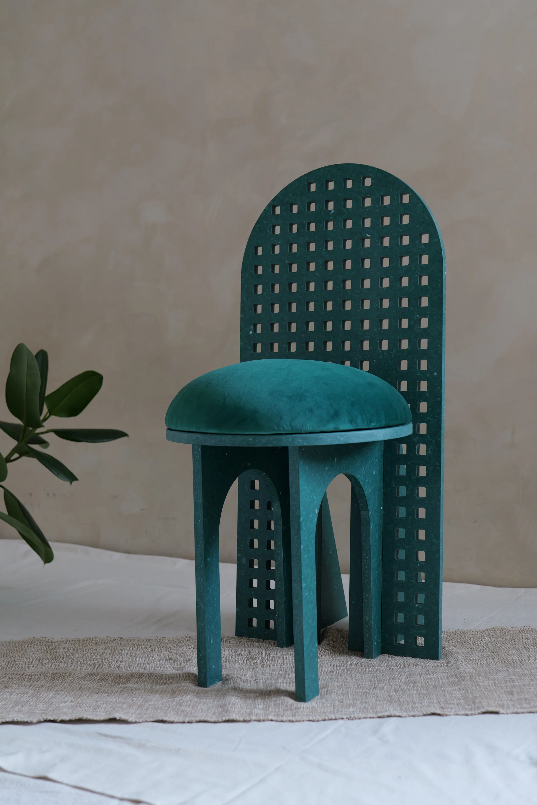 Une chaise bleue en bois avec un coussin rembourré, placée sur un tapis beige, avec une plante verte à gauche et un mur beige en arrière-plan.