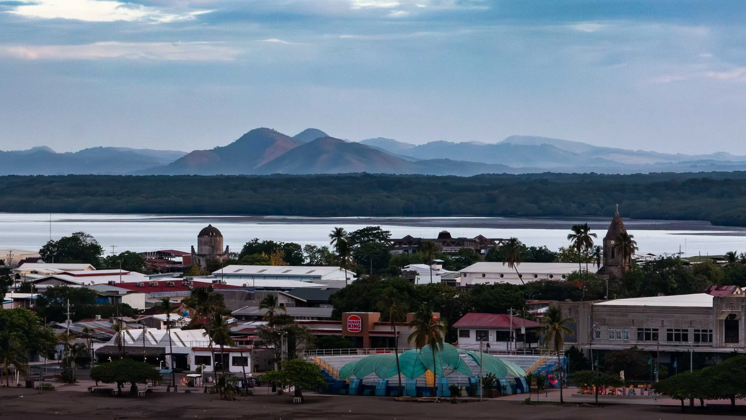 Coastal city of Puntarenas, Costa Rica. Looking across the city to the Gulf of Nicoya and the mountainous Nicoya Peninsula in the background. The green wave-shaped canopy is Plaza de la Artesania. The church tower to the right is Cathedral of Our Lad