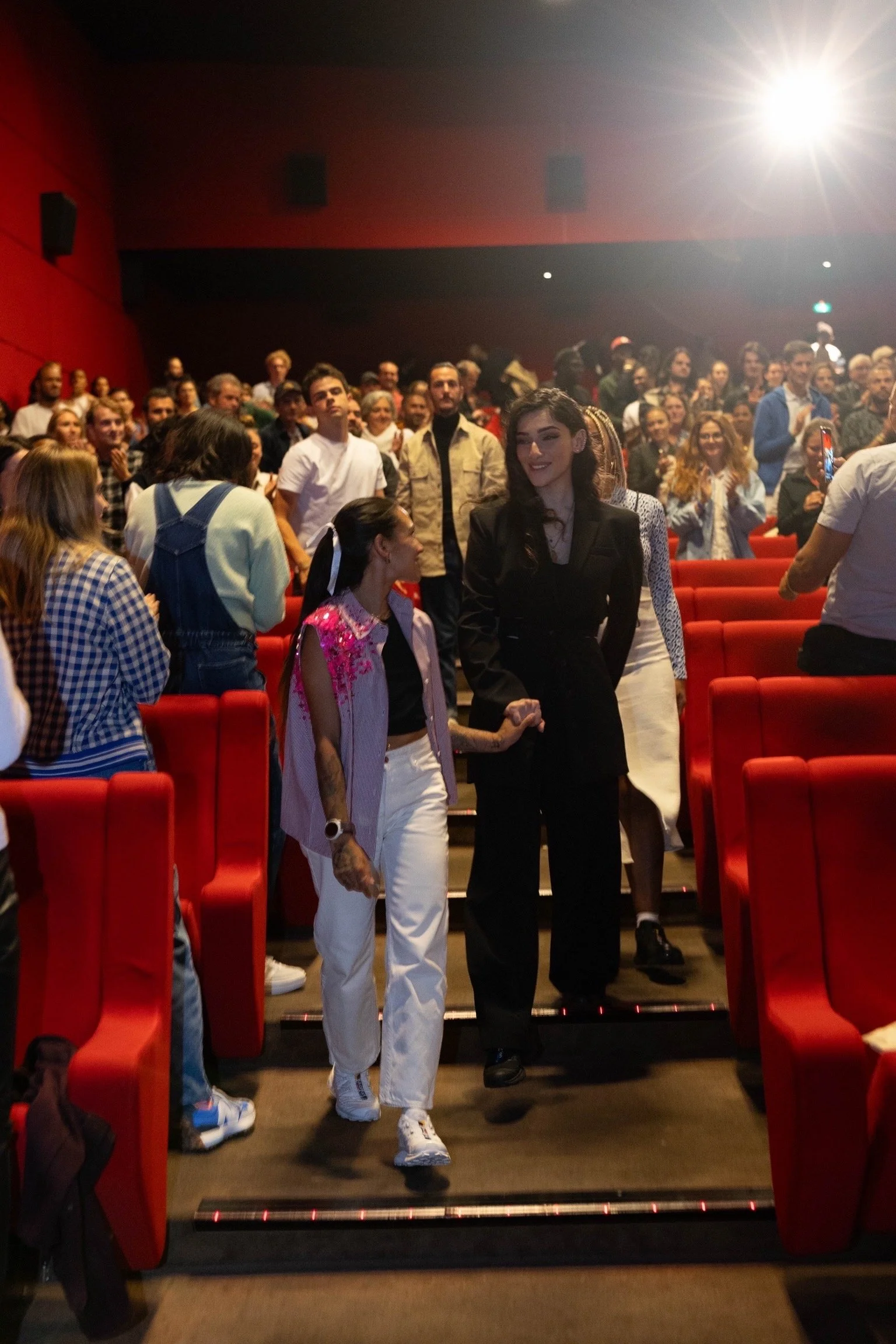 Two women walk down the aisle in a theater, holding hands, with a large audience behind them.