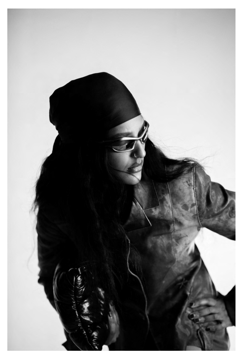 Black and white portrait of a woman wearing sunglasses, a headscarf, and a leather jacket, looking downward with her hand on her hip.
