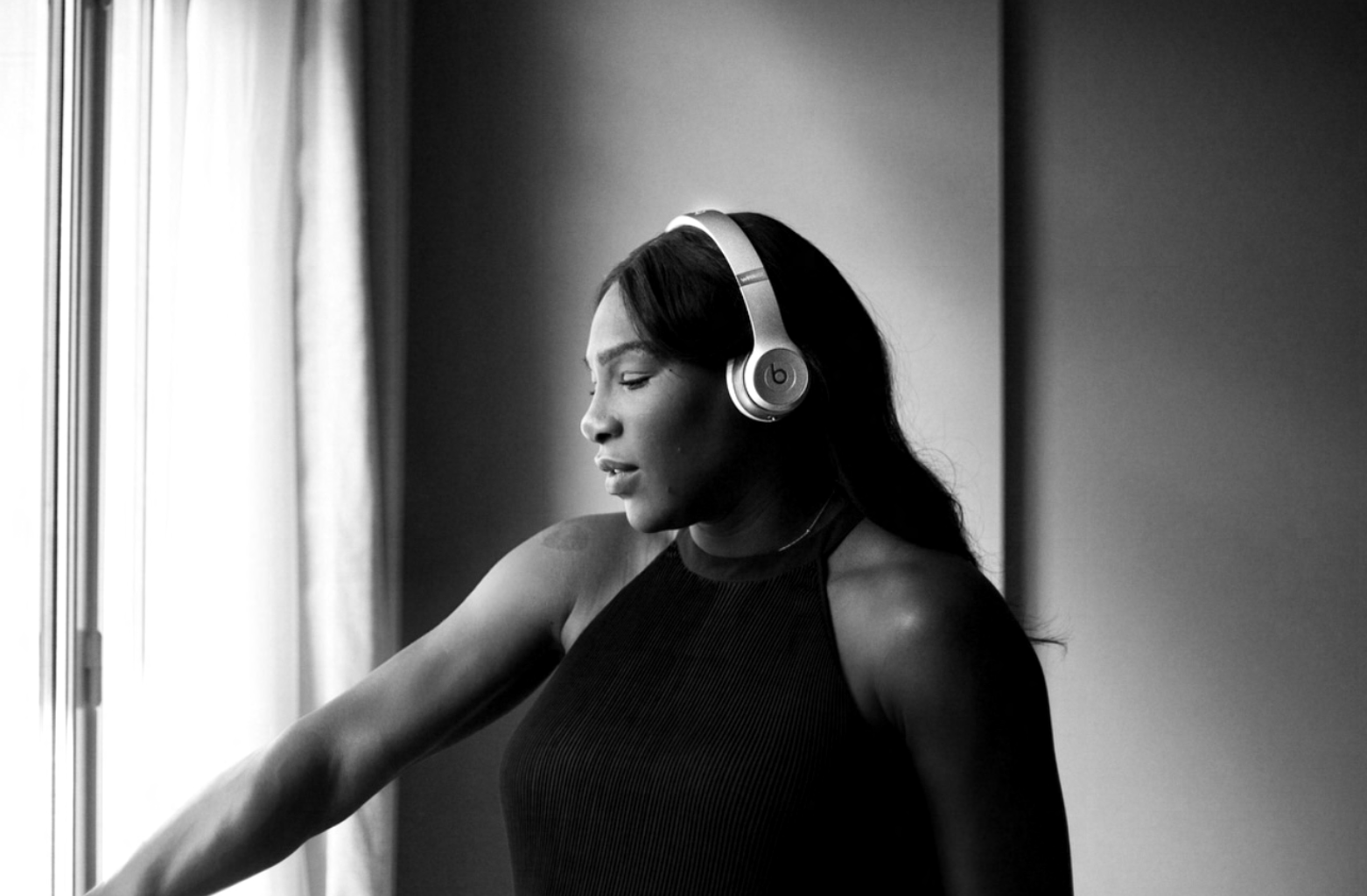 Black and white photo of a woman with long dark hair wearing headphones and a sleeveless top, standing near a window.