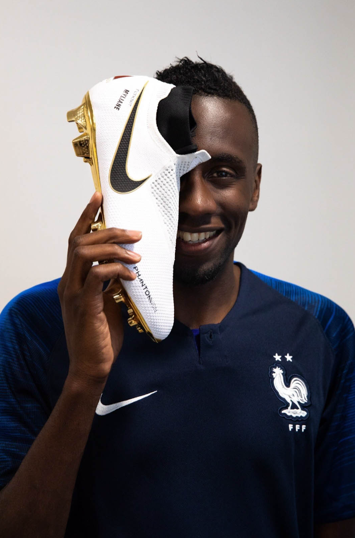 A man in a blue sports jersey holding a Nike soccer cleat up to his face, covering one eye, with a smile.