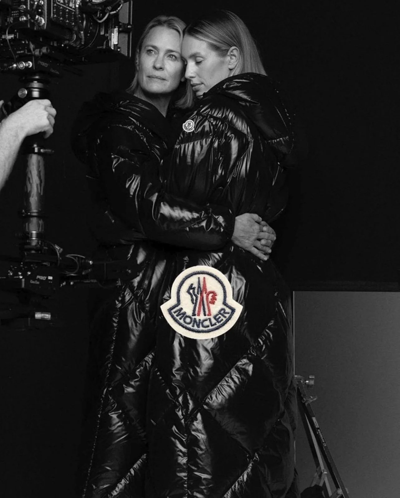 Two women in shiny black Moncler jackets hugging each other in a studio with black background, next to a camera and a Moncler patch on the jackets.