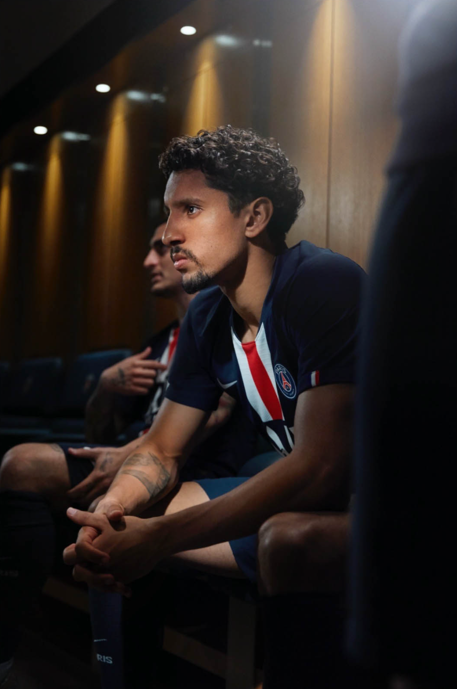 A young man with curly hair and a goatee, wearing a Paris Saint-Germain football jersey, sitting attentively in a dimly lit locker room or team area.