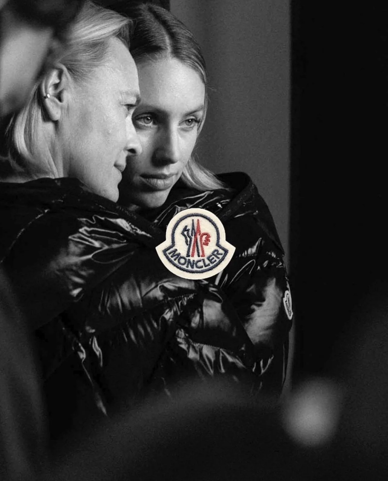 Black and white photo of two women close together, one resting her head on the other's shoulder. The woman on the right is wearing a shiny black jacket with a Moncler logo patch.