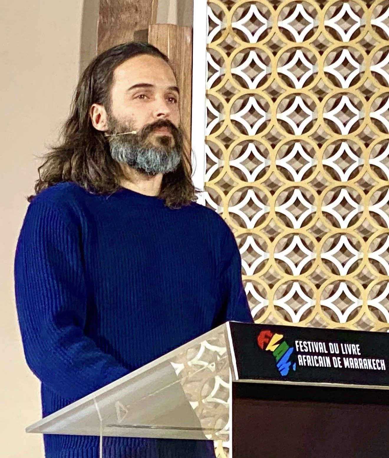 A man with shoulder-length hair and a beard, wearing a blue sweater, stands at a podium during the Festival du Livre Africain de Marrakech.