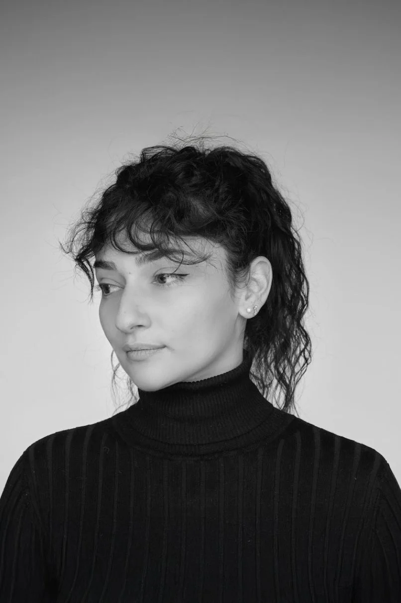 Black and white portrait of a woman looking to her left, wearing a black turtleneck with a textured pattern, dark curly hair, and small earrings.