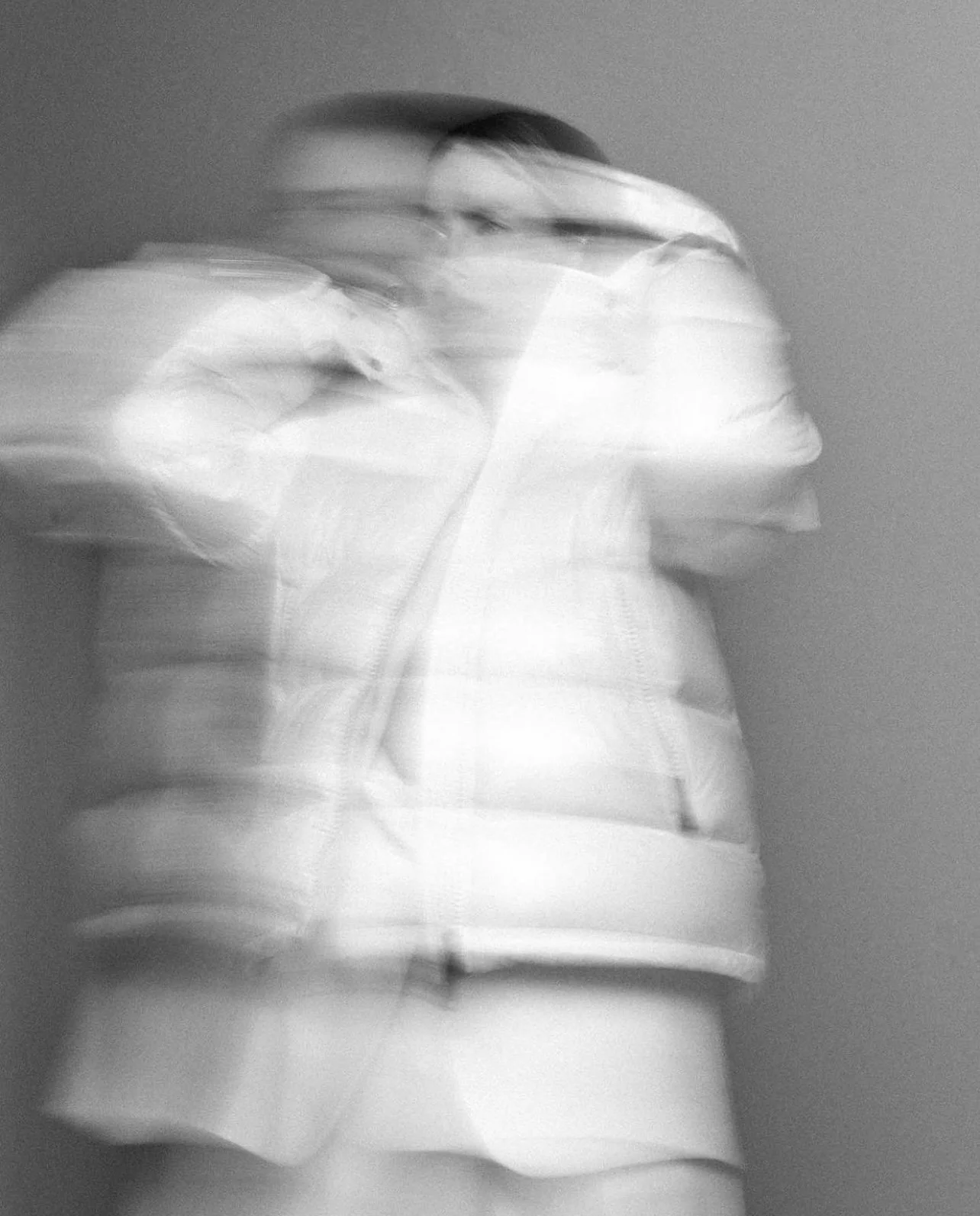Black and white photo of a person with a blurred, motion effect, creating multiple overlapping images of the person's face and upper body, wearing a puffy jacket.