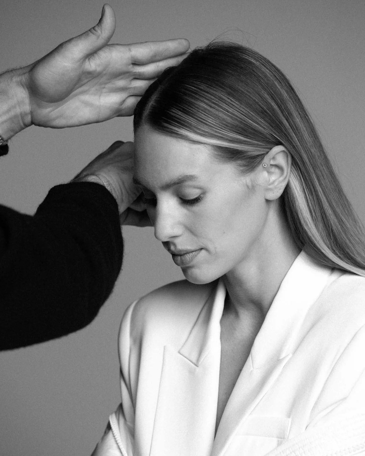 A woman with straight, sleek hair wearing a white blazer, with section of hair being styled by a person with a hand above her head.