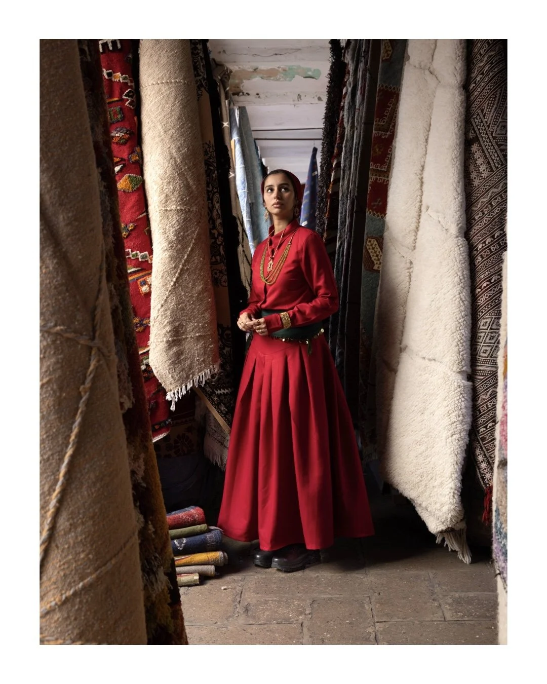 A woman in a red traditional dress with gold jewelry stands amidst hanging carpets in a rug shop.