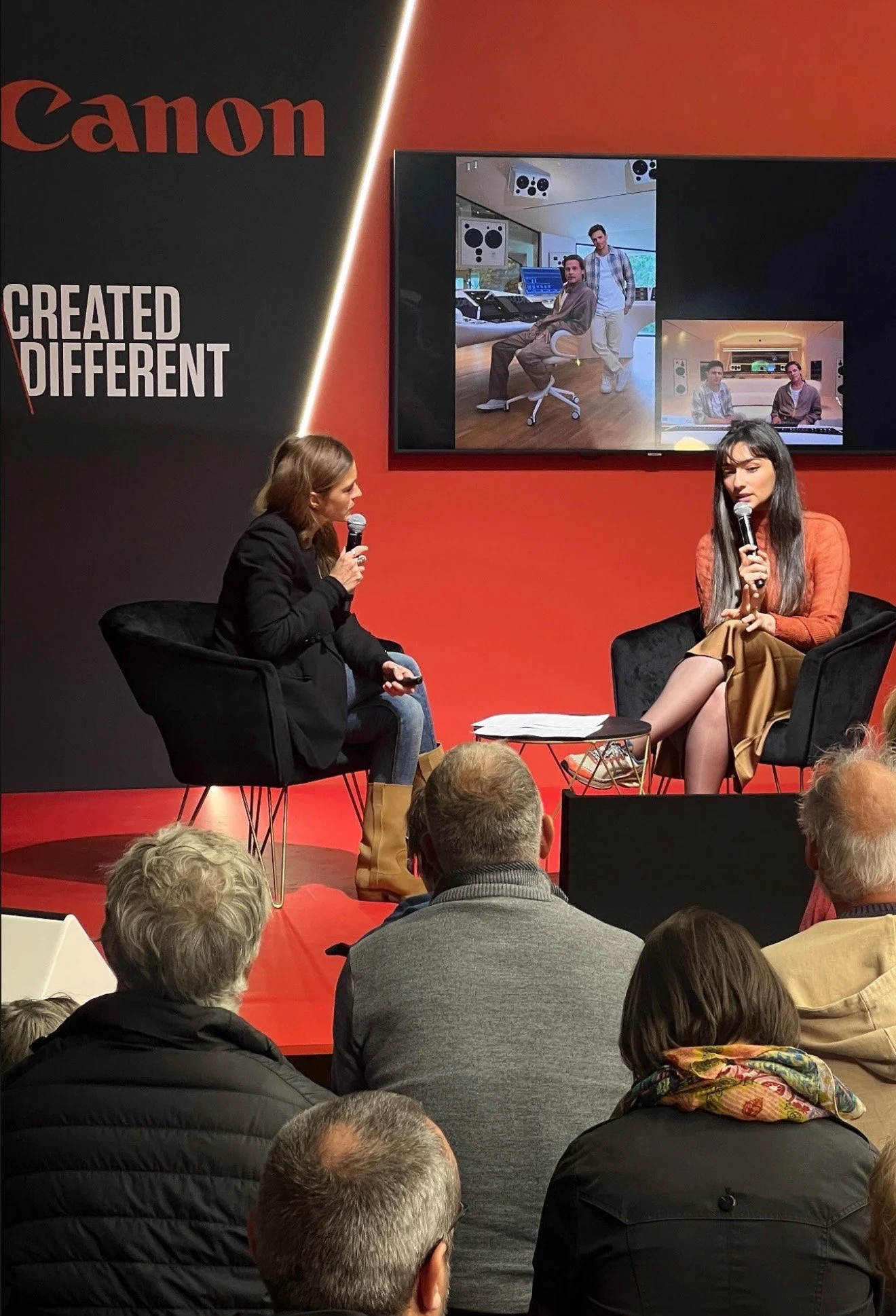 Two women are seated on stage in black armchairs, engaged in a discussion during a presentation or interview, with an audience watching. A large screen behind them displays images of a music studio with two men working.