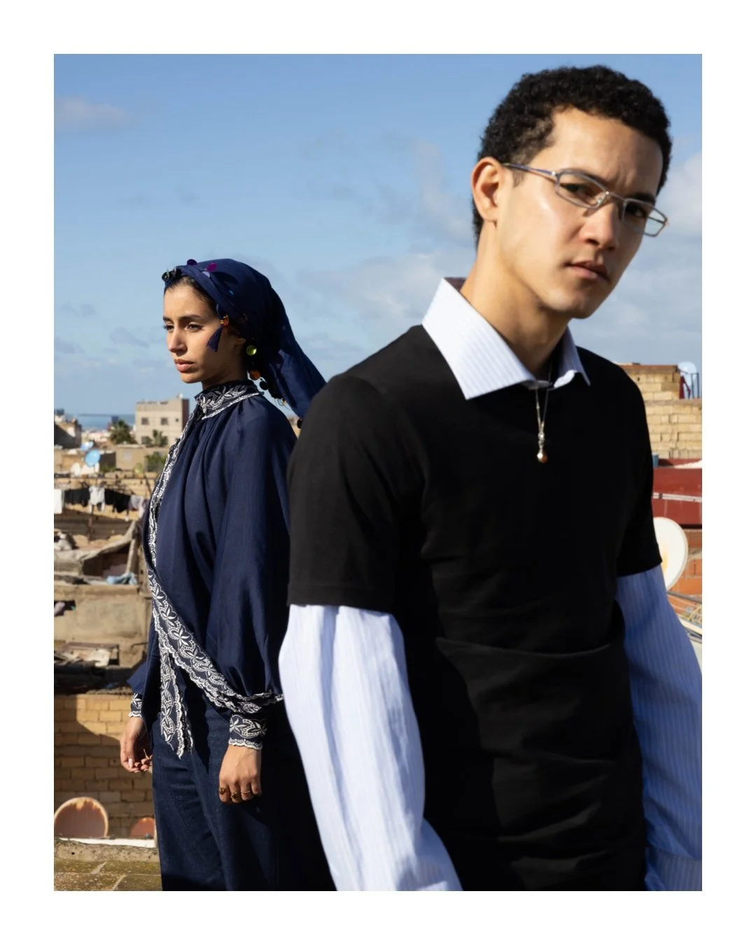A young man and woman standing outdoors on a rooftop against a blue sky with clouds, wearing stylish clothing and glasses.