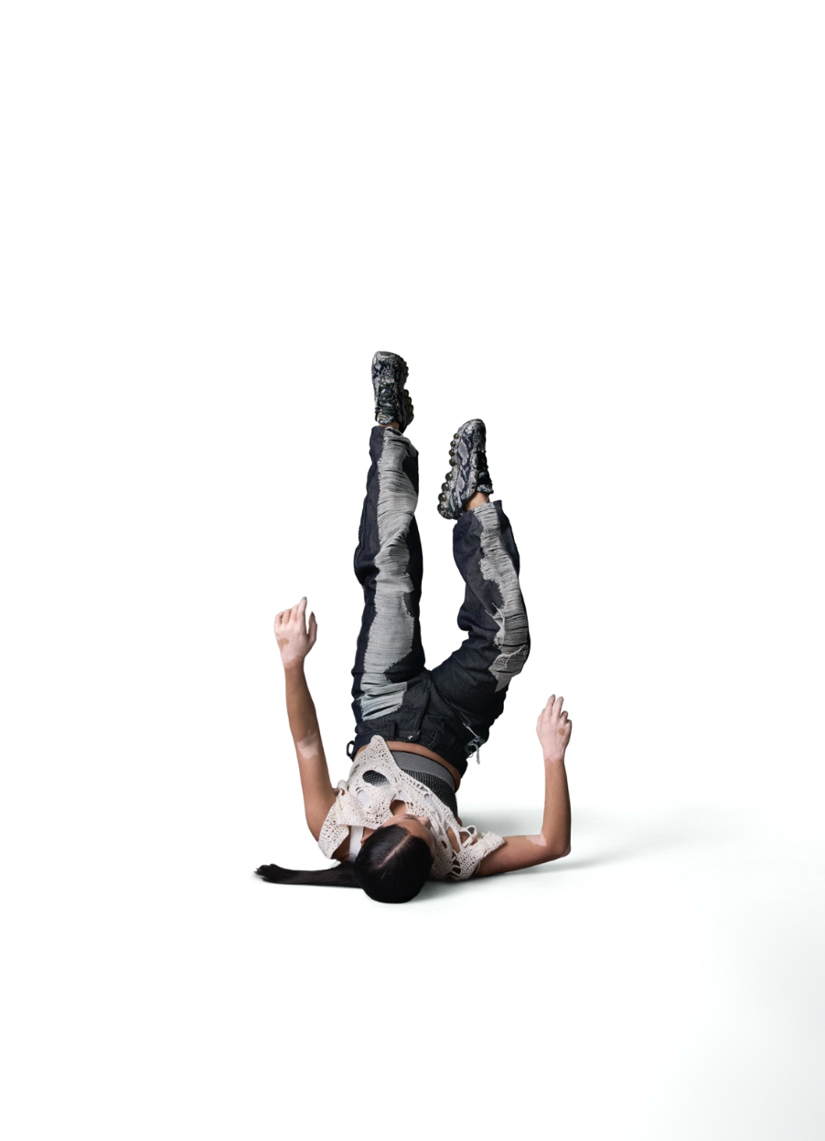 A person lying on their back with their legs raised upright, wearing black and gray camouflage-patterned clothing and black sneakers, against a white background.