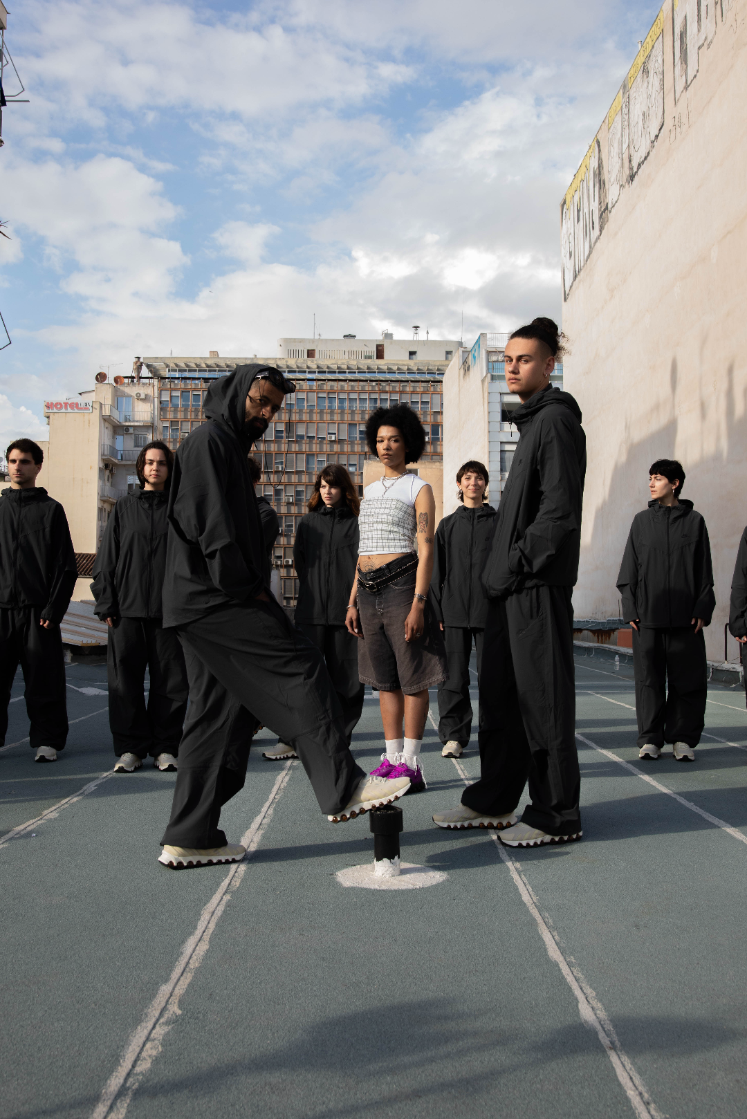 A group of people standing on a rooftop, with one person balancing on a small black post while the others watch. The group includes both men and women, with some dressed in black hoodies and sweatpants, and one woman in casual clothing with purple sn
