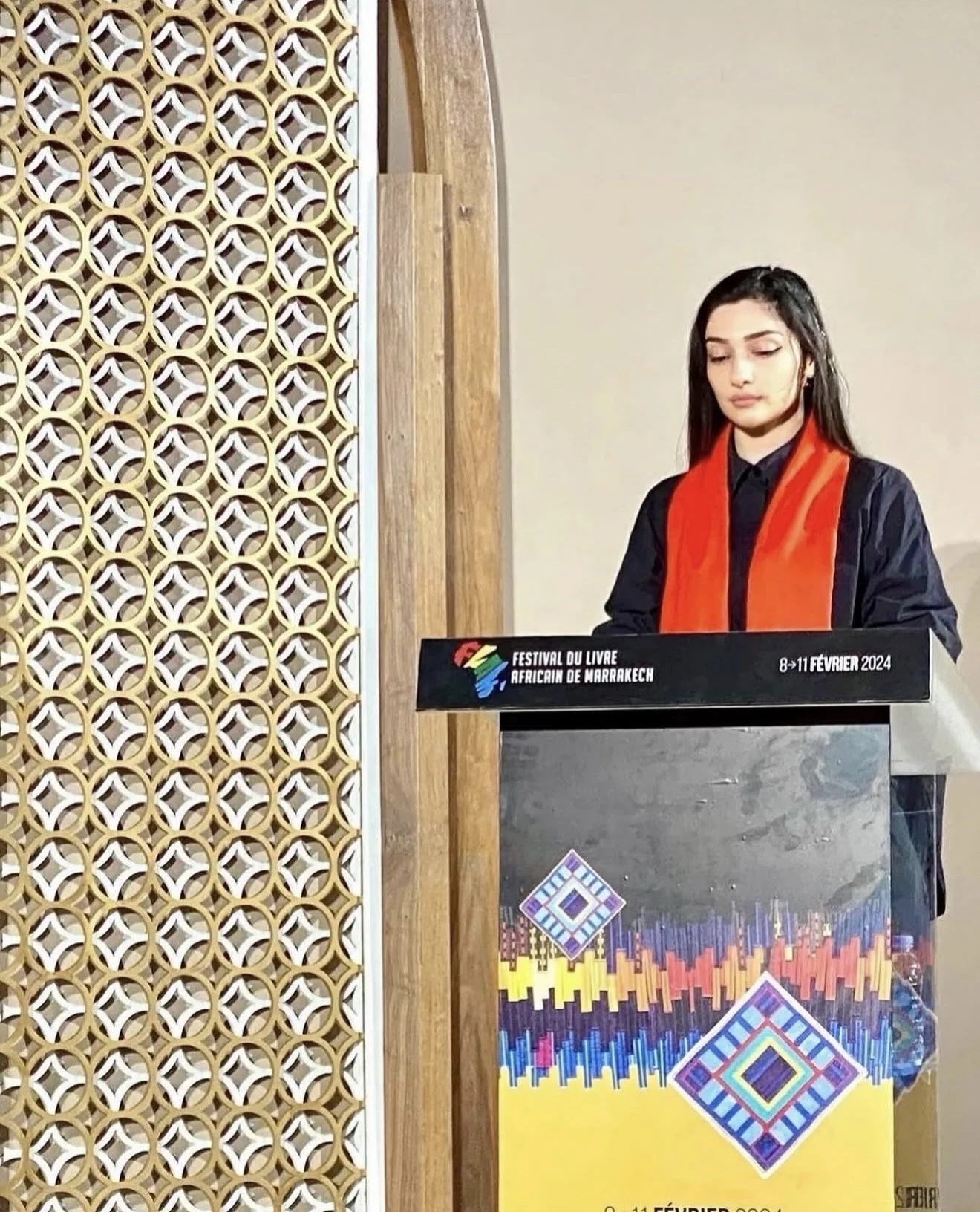 A young woman with long dark hair, wearing a black jacket and a bright orange scarf, stands behind a podium at the African Book Festival in Marrakech, February 8-11, 2024. The podium features colorful geometric artwork, and there is a decorative wooden panel with a circular pattern to the left of her.