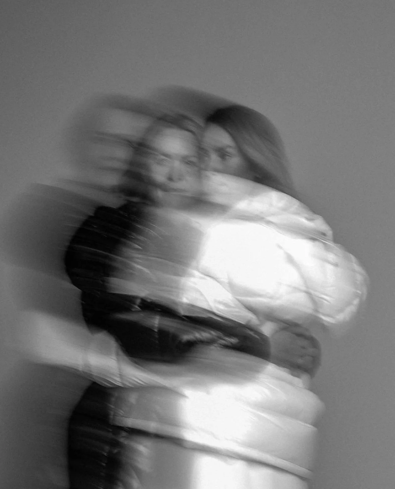 A blurred black and white photo of two women hugging, one wearing glasses and a dark jacket, the other with long hair and a light-colored shirt, against a plain background.