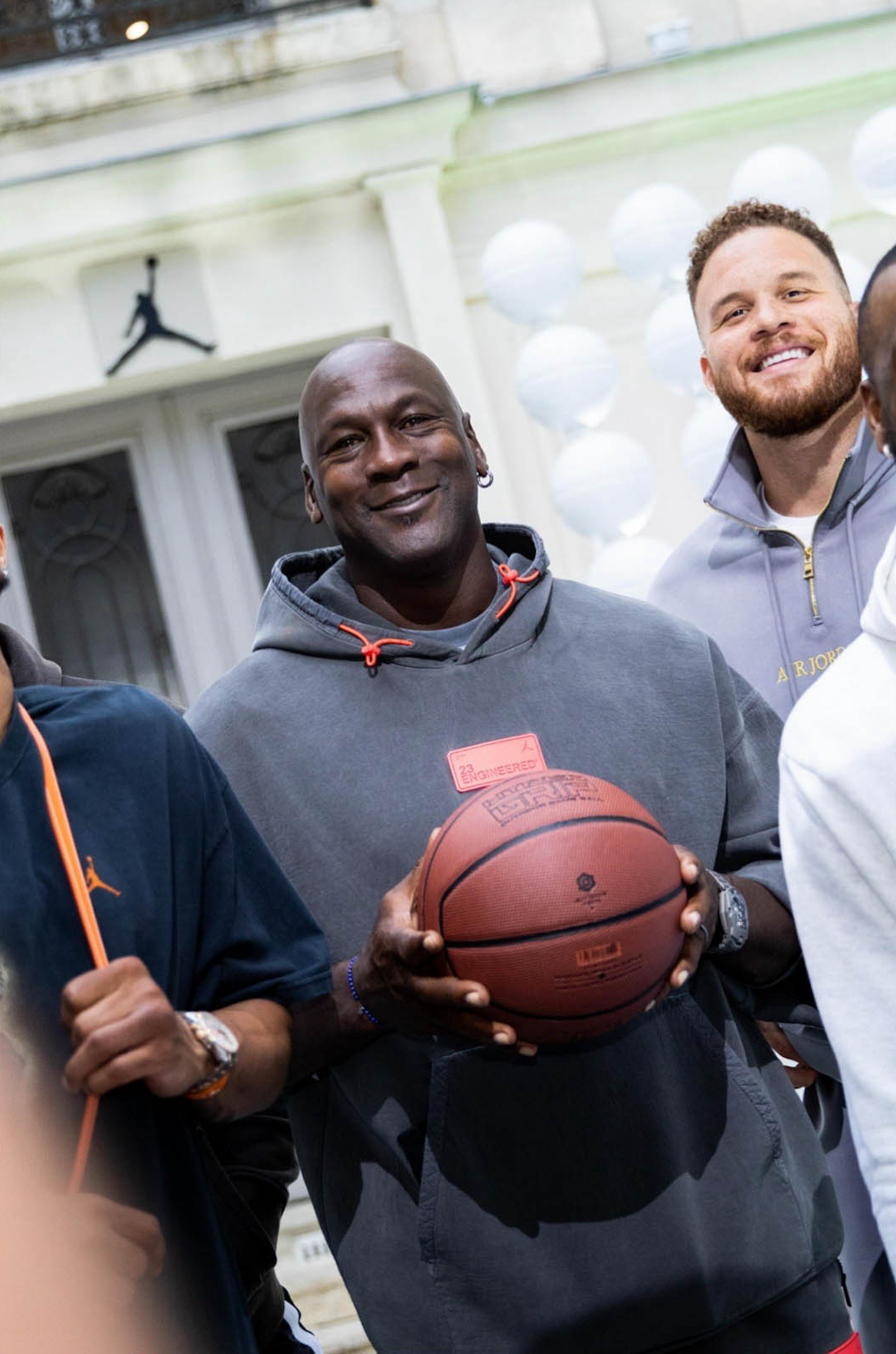A group of men, with one in the center holding a basketball, standing outside a building with a porch and white balloons in the background.