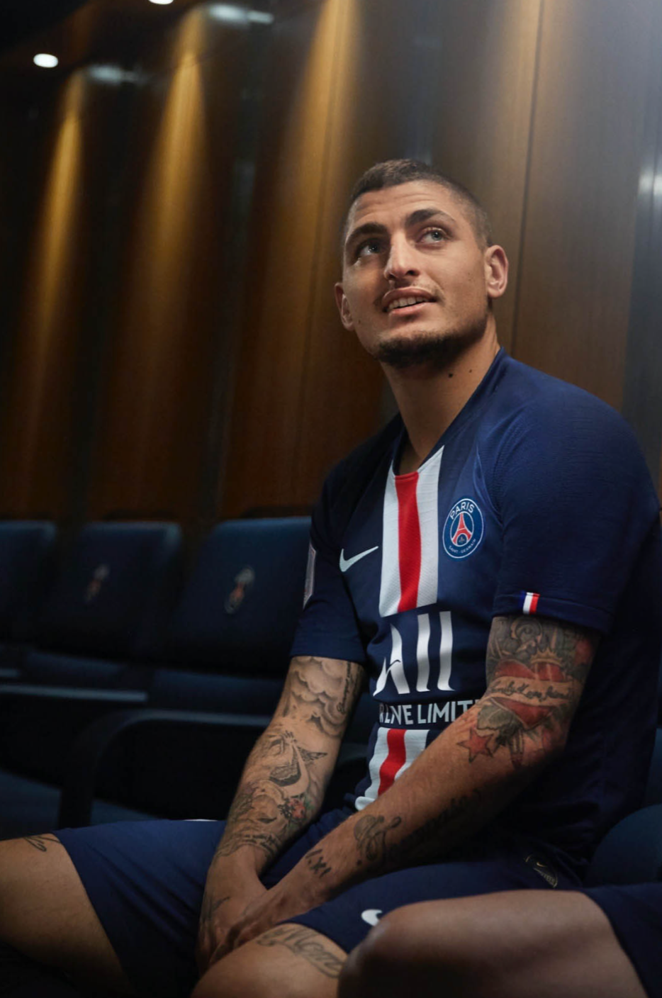 A young man with short hair, tattoos on his arms, and a slight beard, sitting on a bench inside a room with wooden walls, wearing a Paris Saint-Germain football jersey.