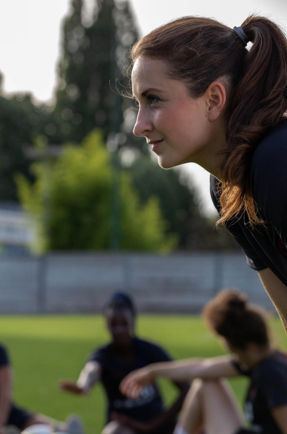 A young woman with brown hair tied in a ponytail leaning forward. She is outdoors on a grassy field with blurred people sitting and a fence in the background.