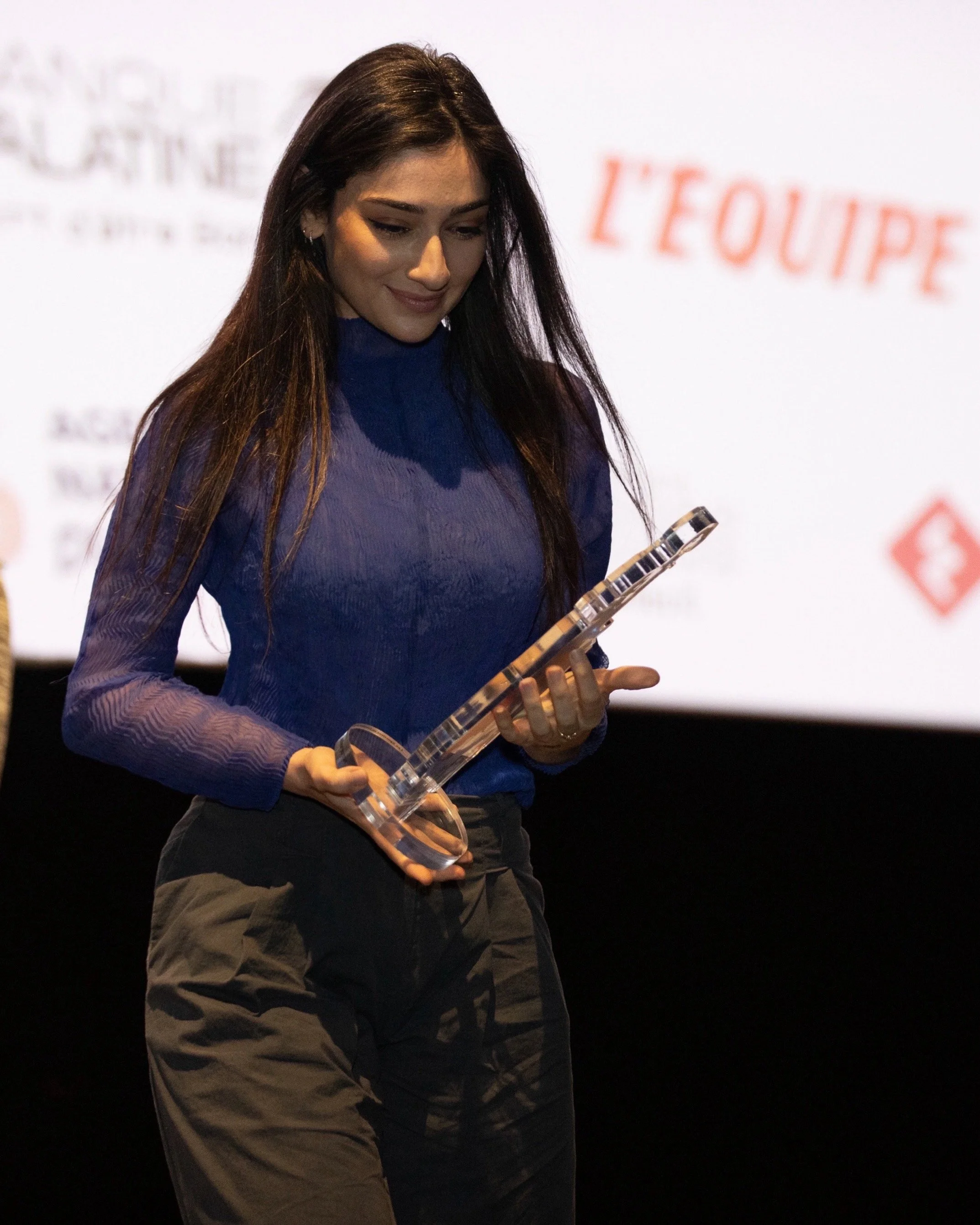A woman with long dark hair, wearing a blue top and brown pants, holding a clear glass award or trophy. She is smiling slightly and looking down at the award in her hands. In the background, there is a blurred white and orange sign with French text.