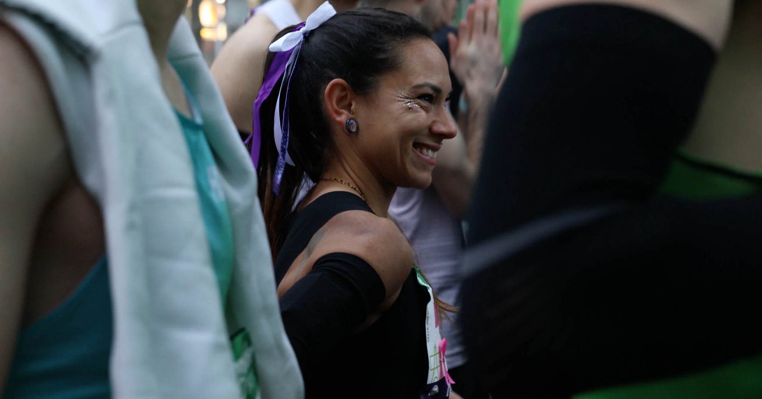 A joyful woman with long dark hair tied with purple and white ribbons, smiling among a crowd, wearing dark clothing and earrings, at what appears to be a public event.