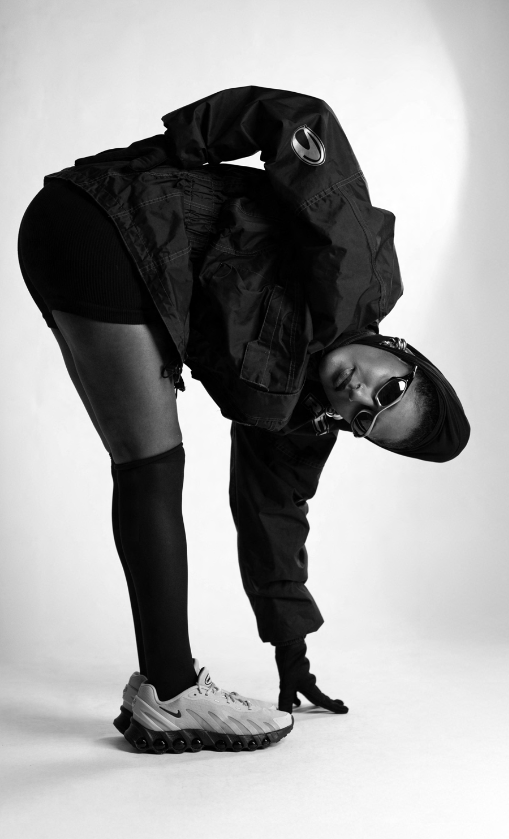 A person dressed in athletic wear, including Nike sneakers, leggings, and a jacket, with sunglasses and a beanie, bending over with one hand on the ground.