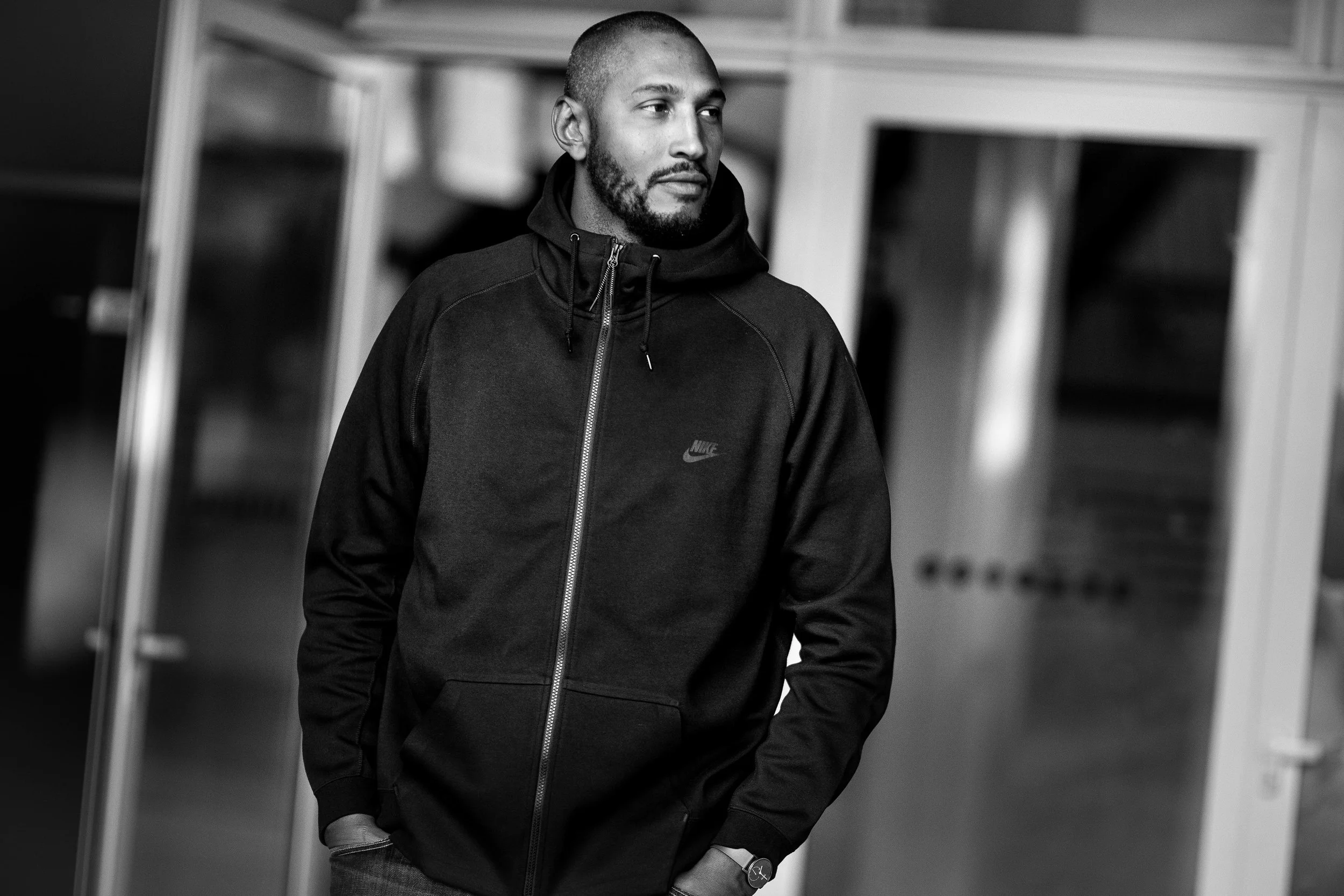 A man in a black Nike hoodie walks through a hallway with glass doors, gazing to the side in a serious expression.